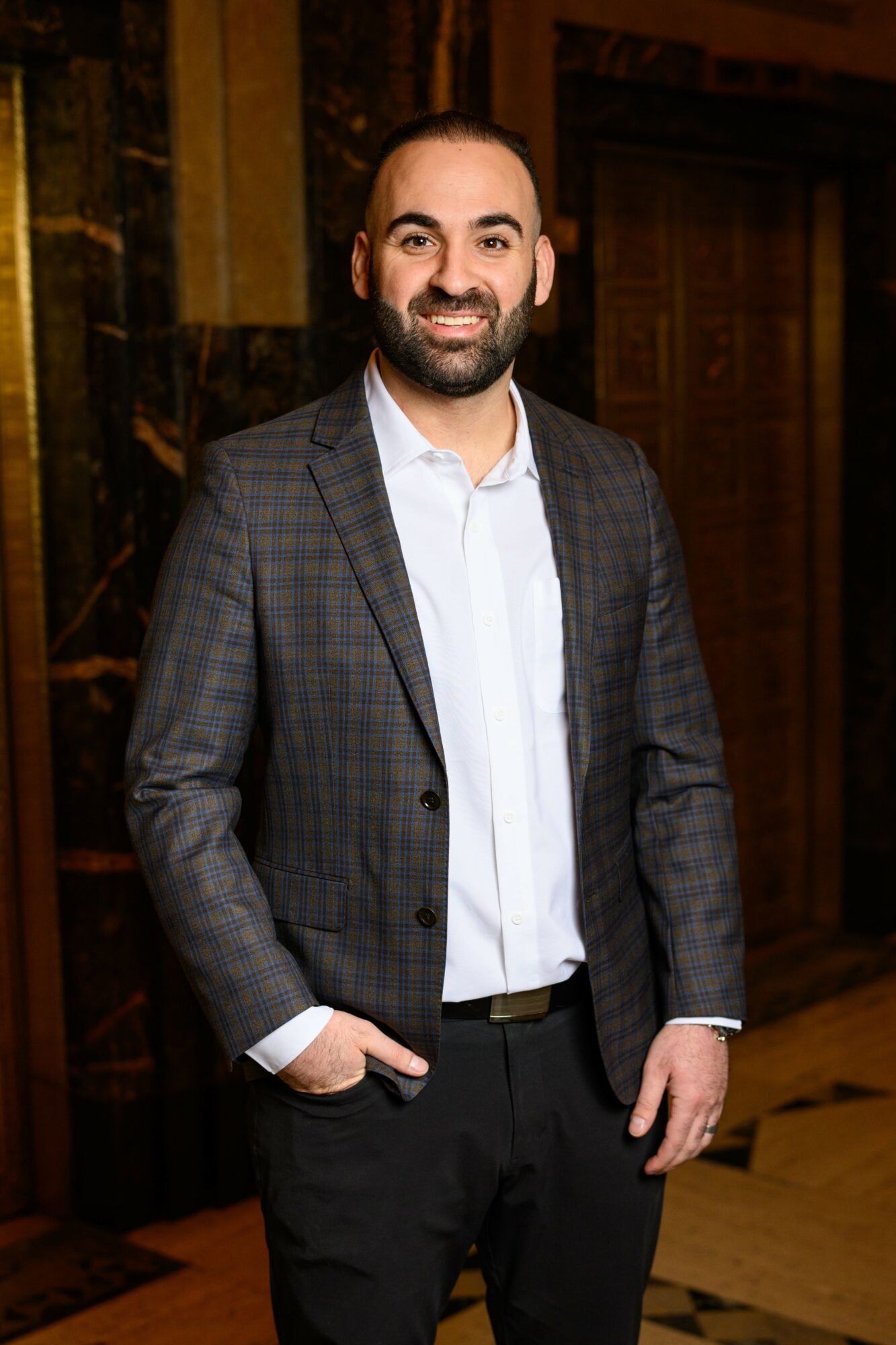 Man with a beard in a suit and white shirt smiling indoors, hands in pockets.