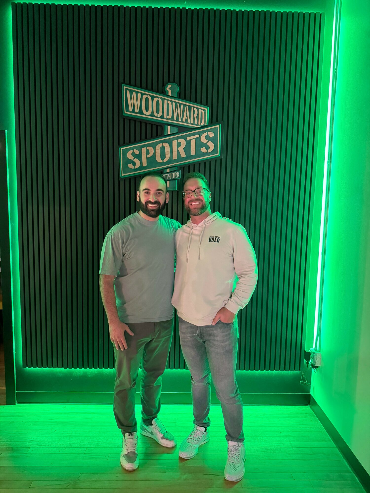 Two people standing in front of a sports sign, illuminated by green lighting, with a textured wall background.