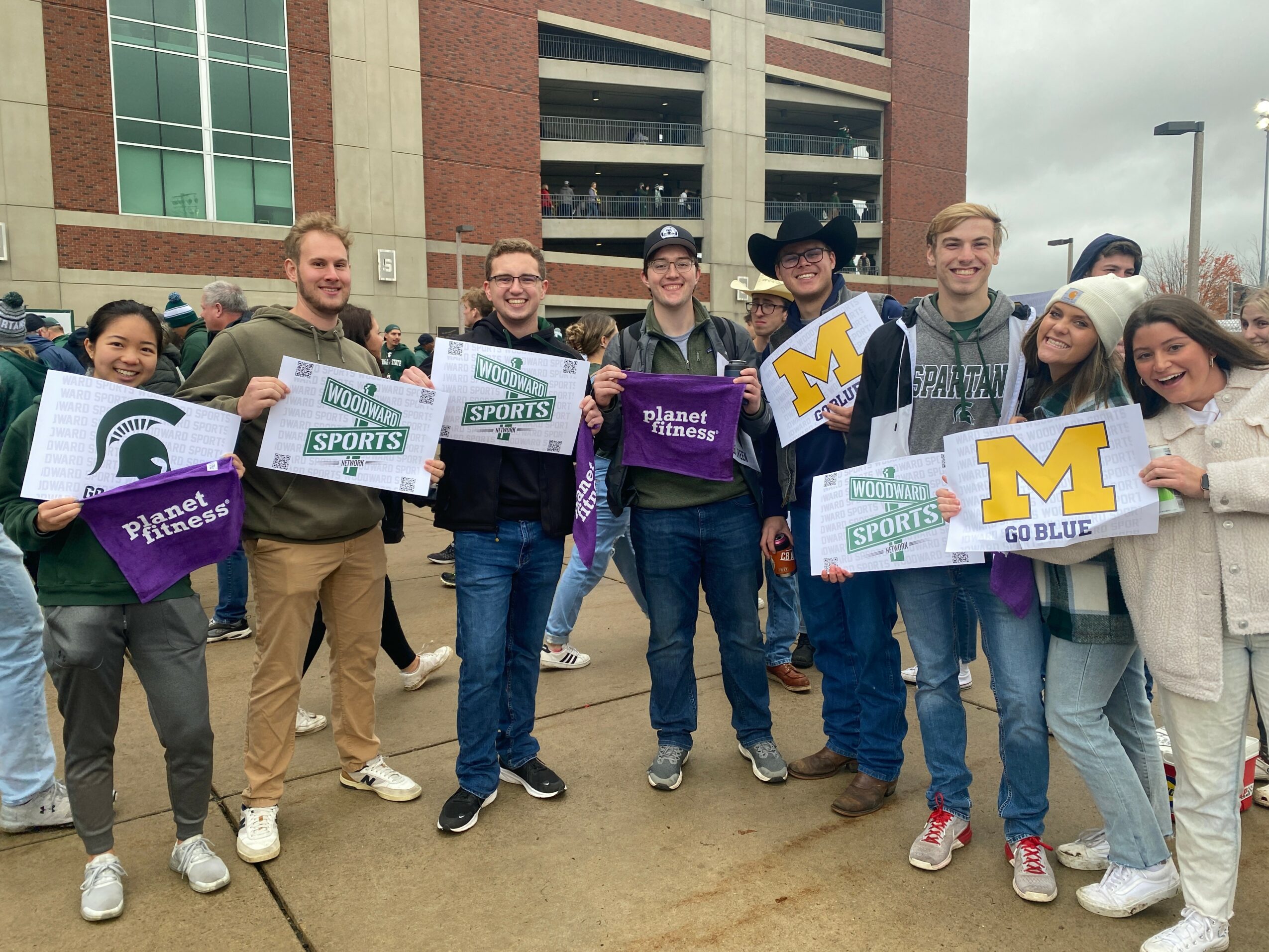 Group of people holding signs and banners outside a building, smiling, with cloudy sky overhead.