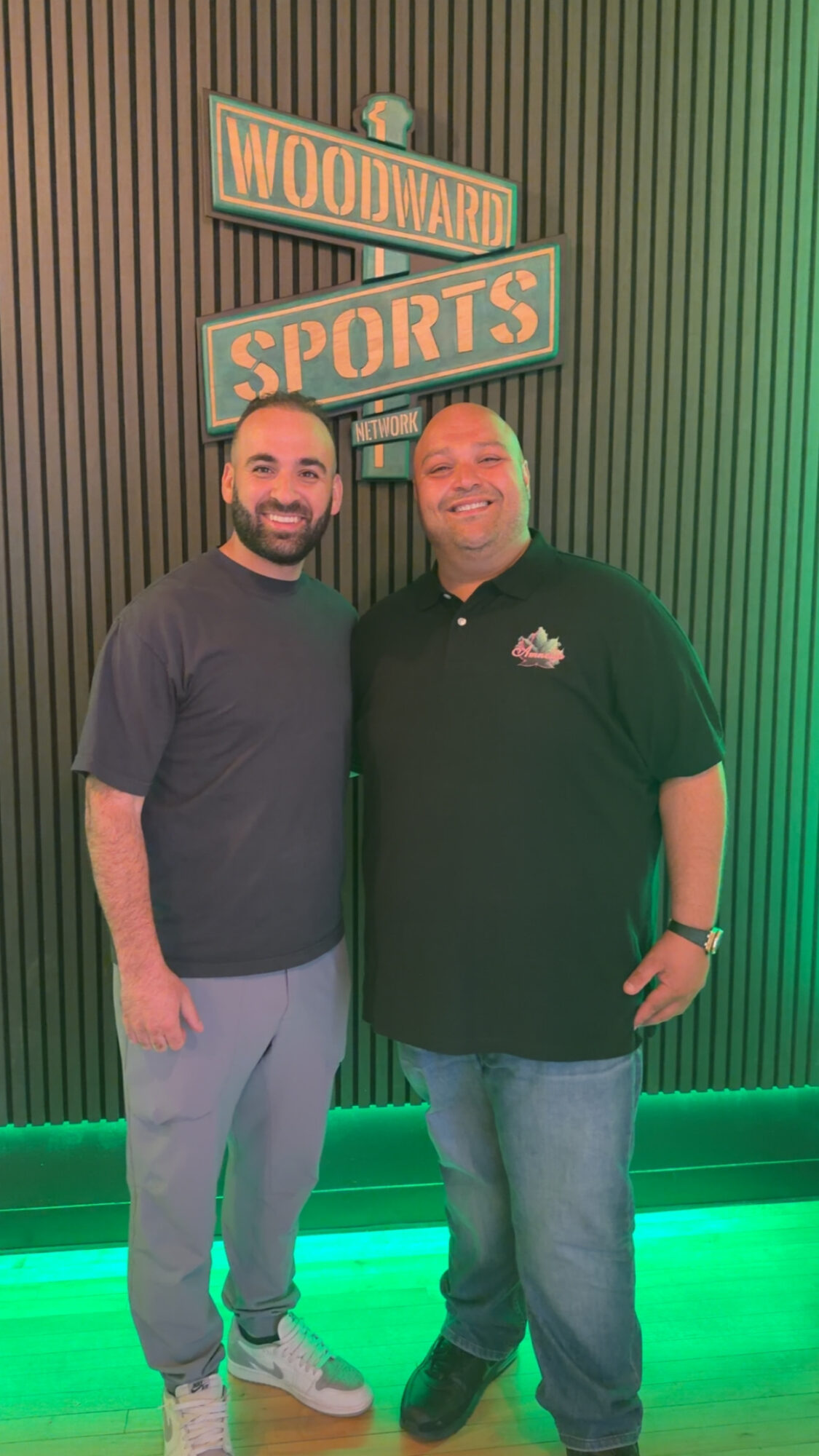 Two men stand together in front of a wall with Woodward Sports signs, smiling, with green lighting on the floor.