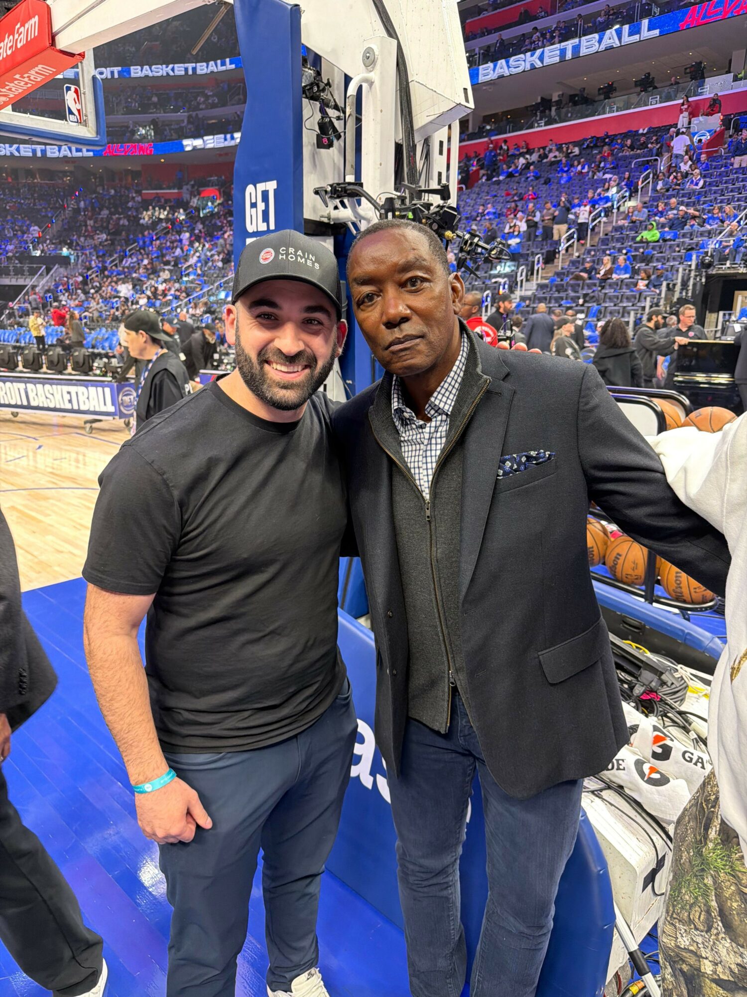 Two men standing together in a sports arena, one in a suit and the other in casual clothing, with a crowd in the background.