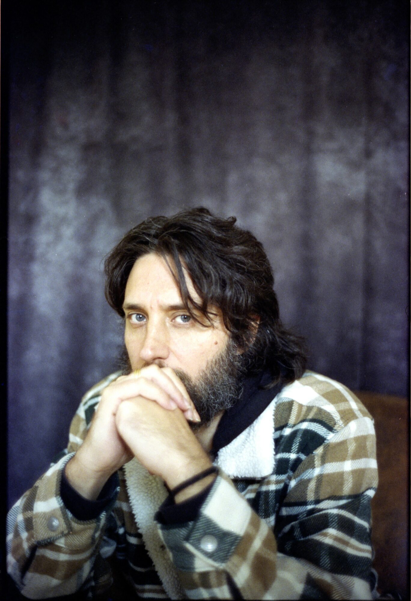 Man with dark hair and beard, wearing a plaid jacket, sitting with hands near face, against a dark background.