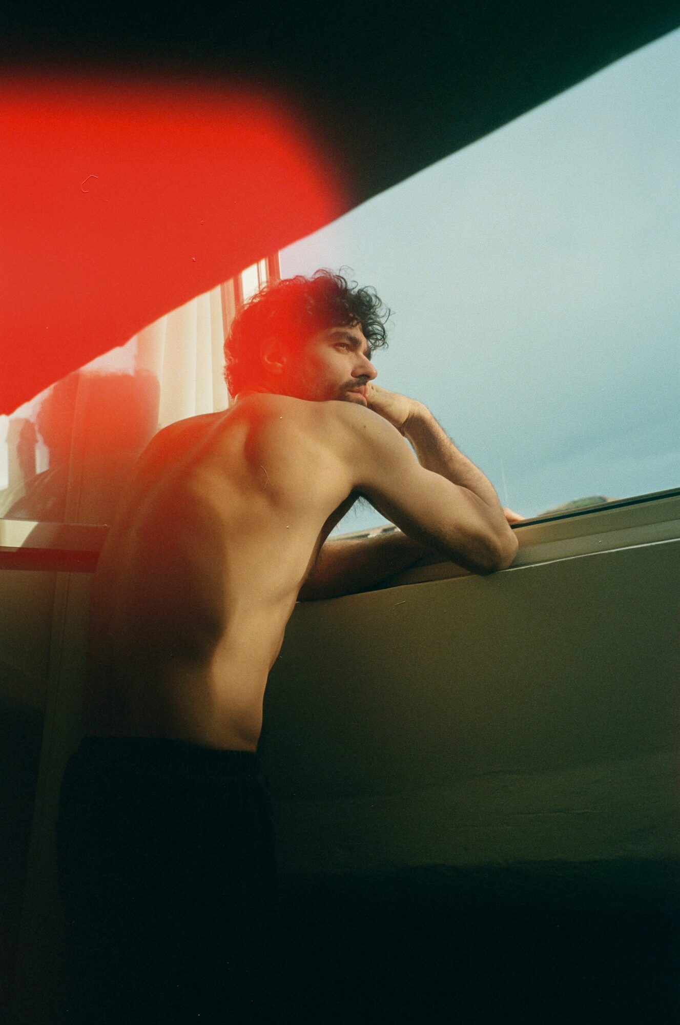 A shirtless person with curly hair leans on a window, looking outside with a thoughtful expression.