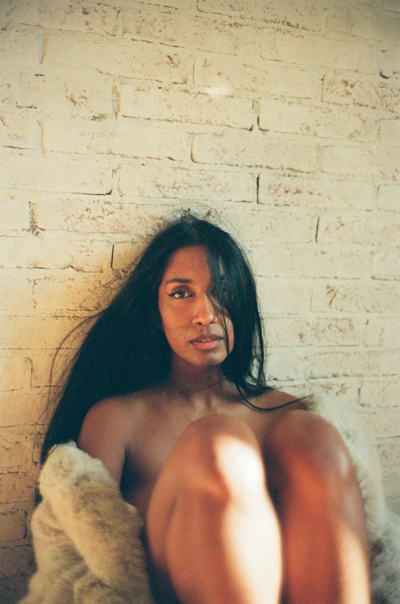 Woman with long dark hair sitting against a white brick wall, looking at the camera, with knees up.