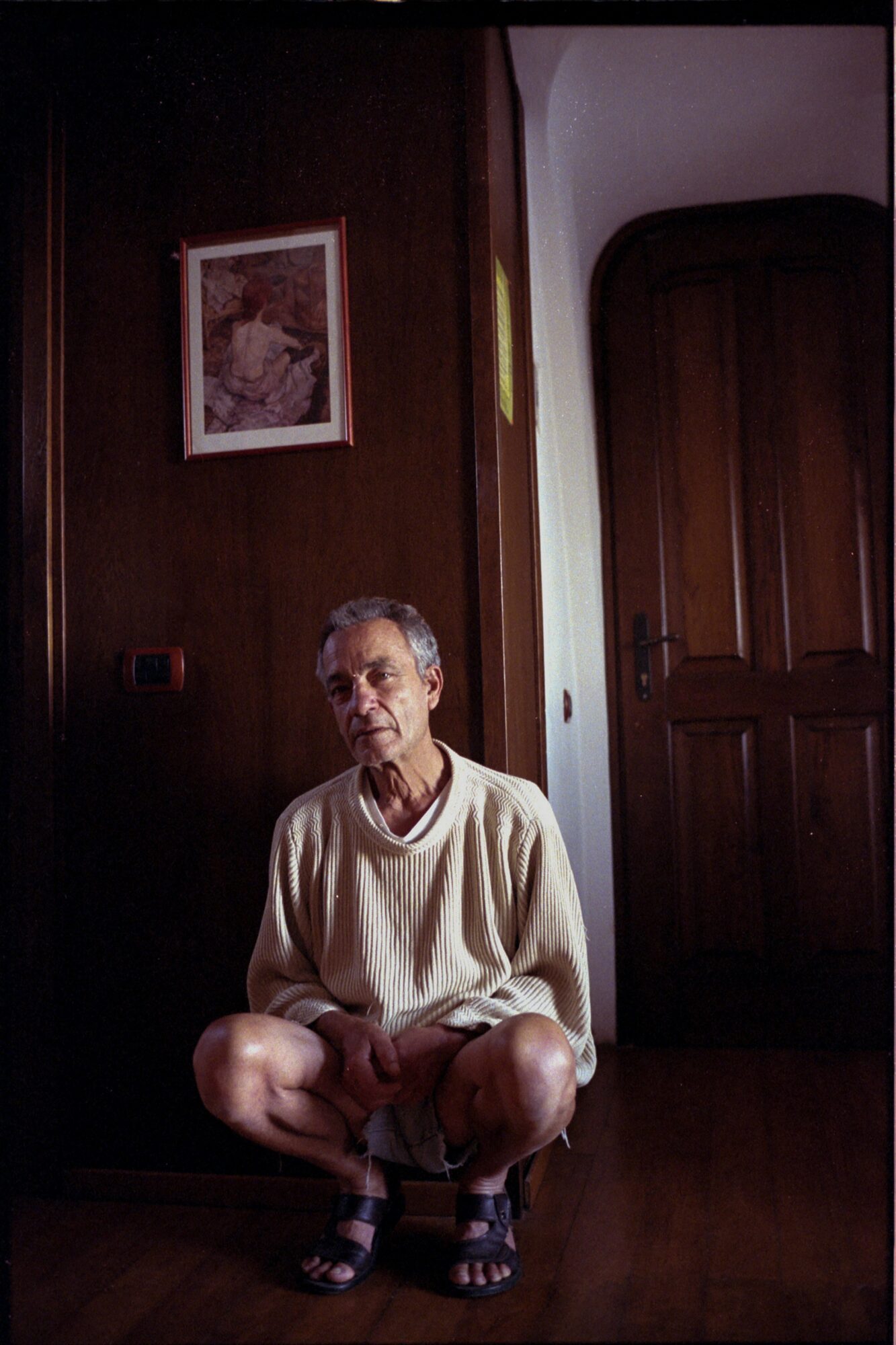 Elderly person crouching indoors, wooden door and framed picture on wall behind, wearing a light sweater and sandals.
