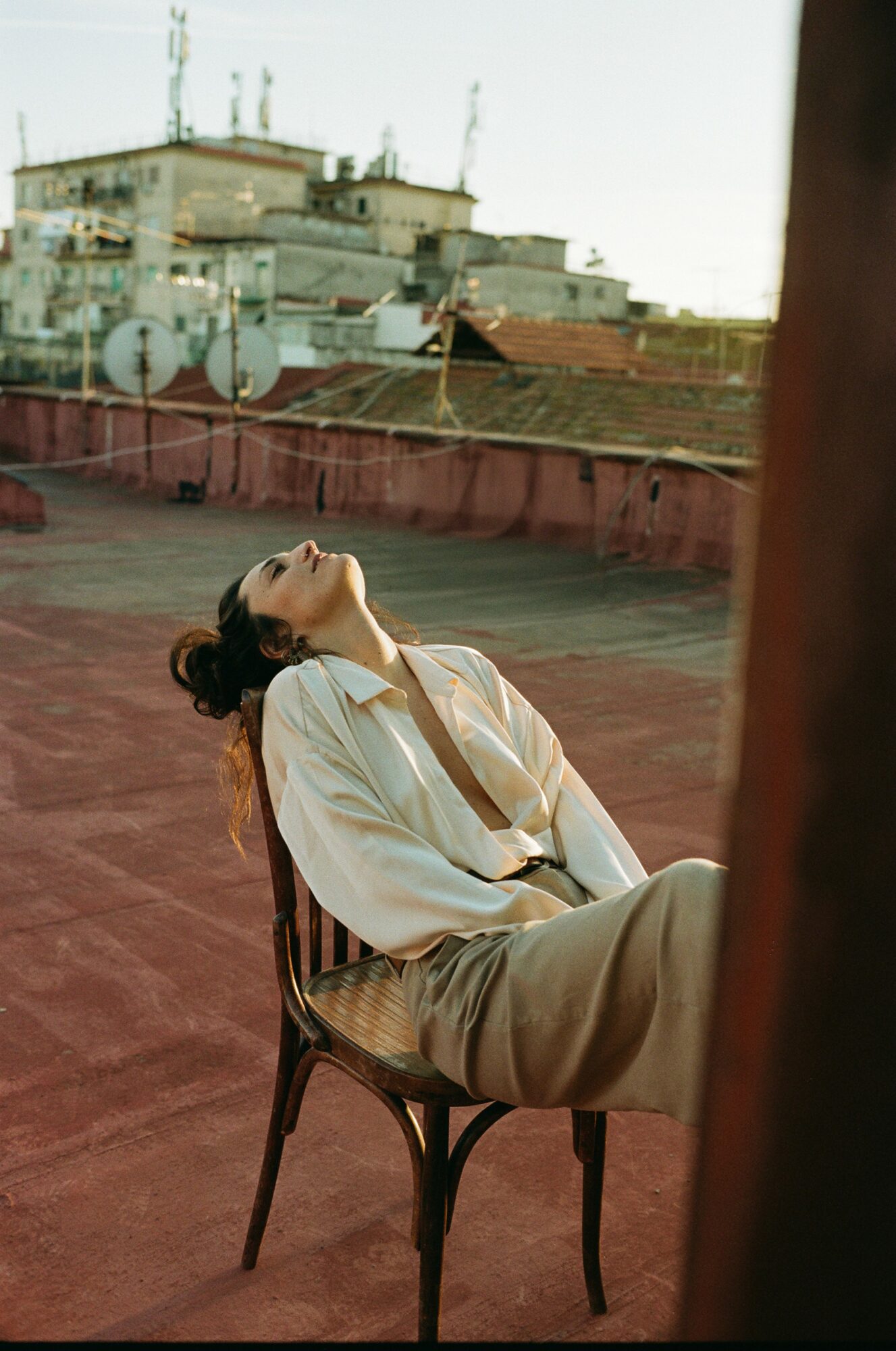 Person sitting on a chair on a rooftop, leaning back with head tilted upward, cityscape in background.