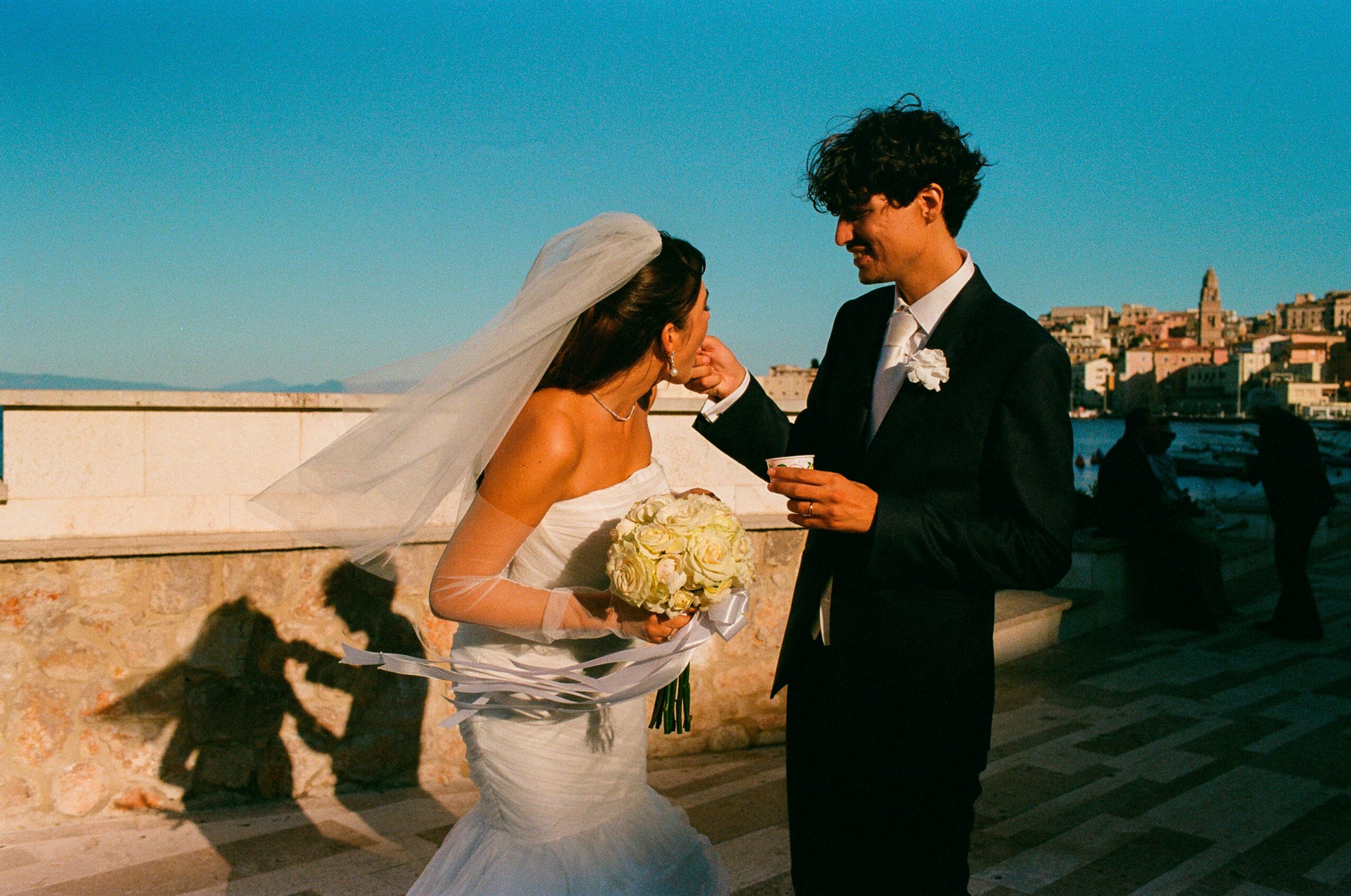 Bride and groom exchanging rings outdoors, bride holding a bouquet, sunny day, cityscape in background.