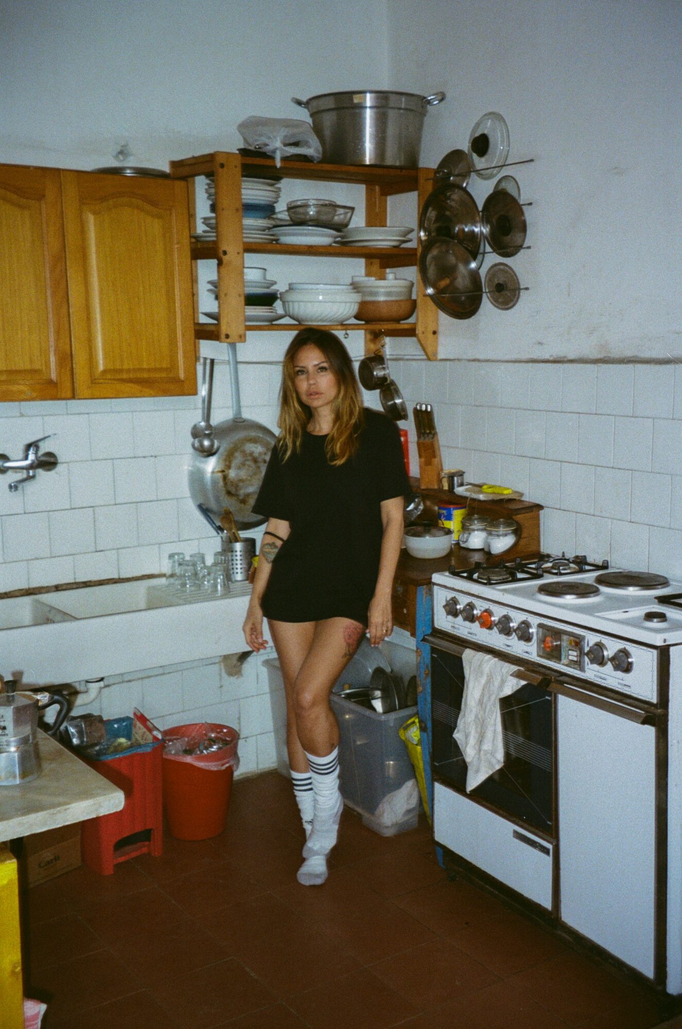 Young woman in black dress and knee-high socks standing in a cluttered kitchen with stove, shelves, and dishes.