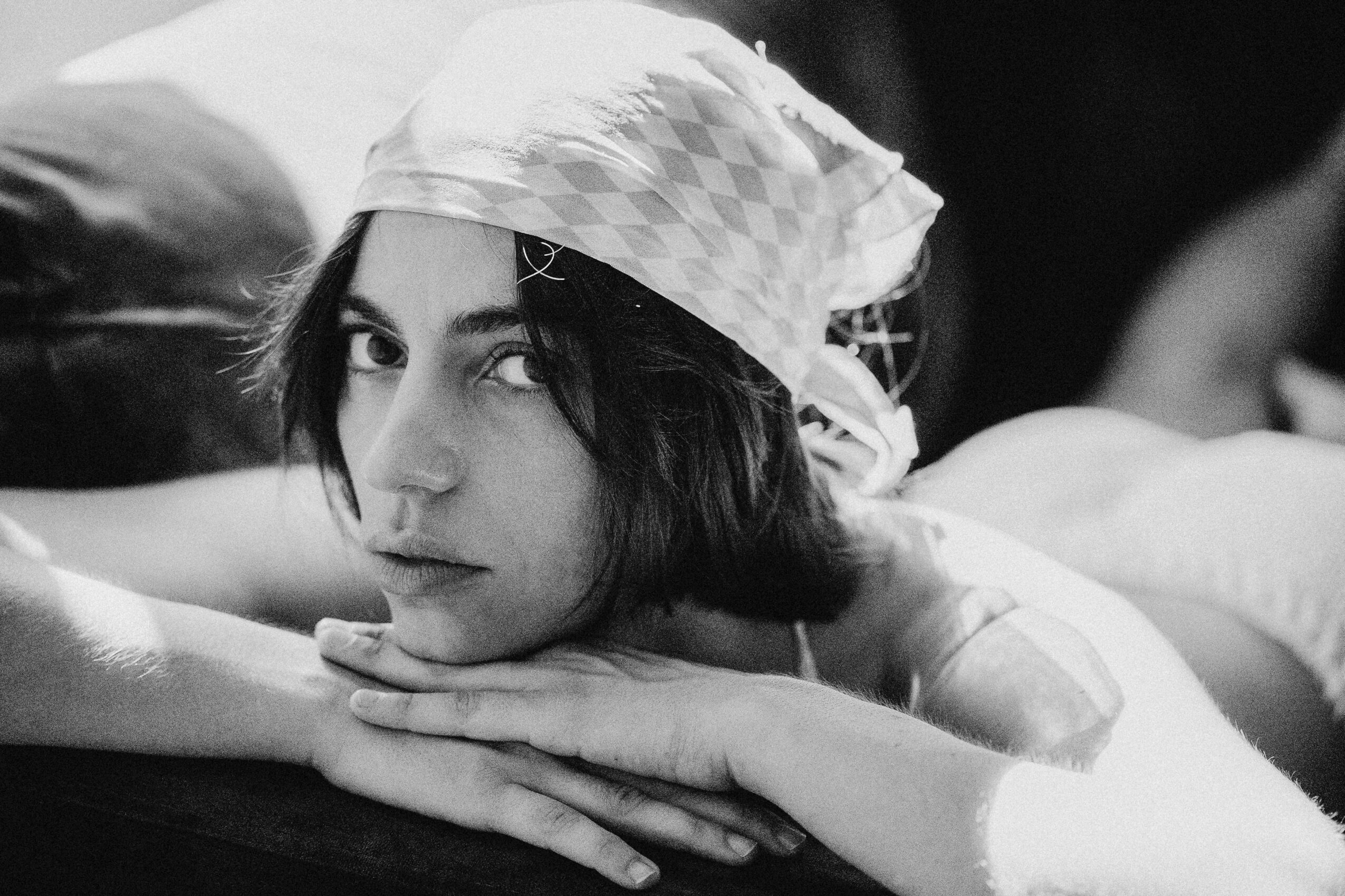 Woman with short dark hair wearing a patterned headscarf, resting her chin on her hands, looking at the camera.