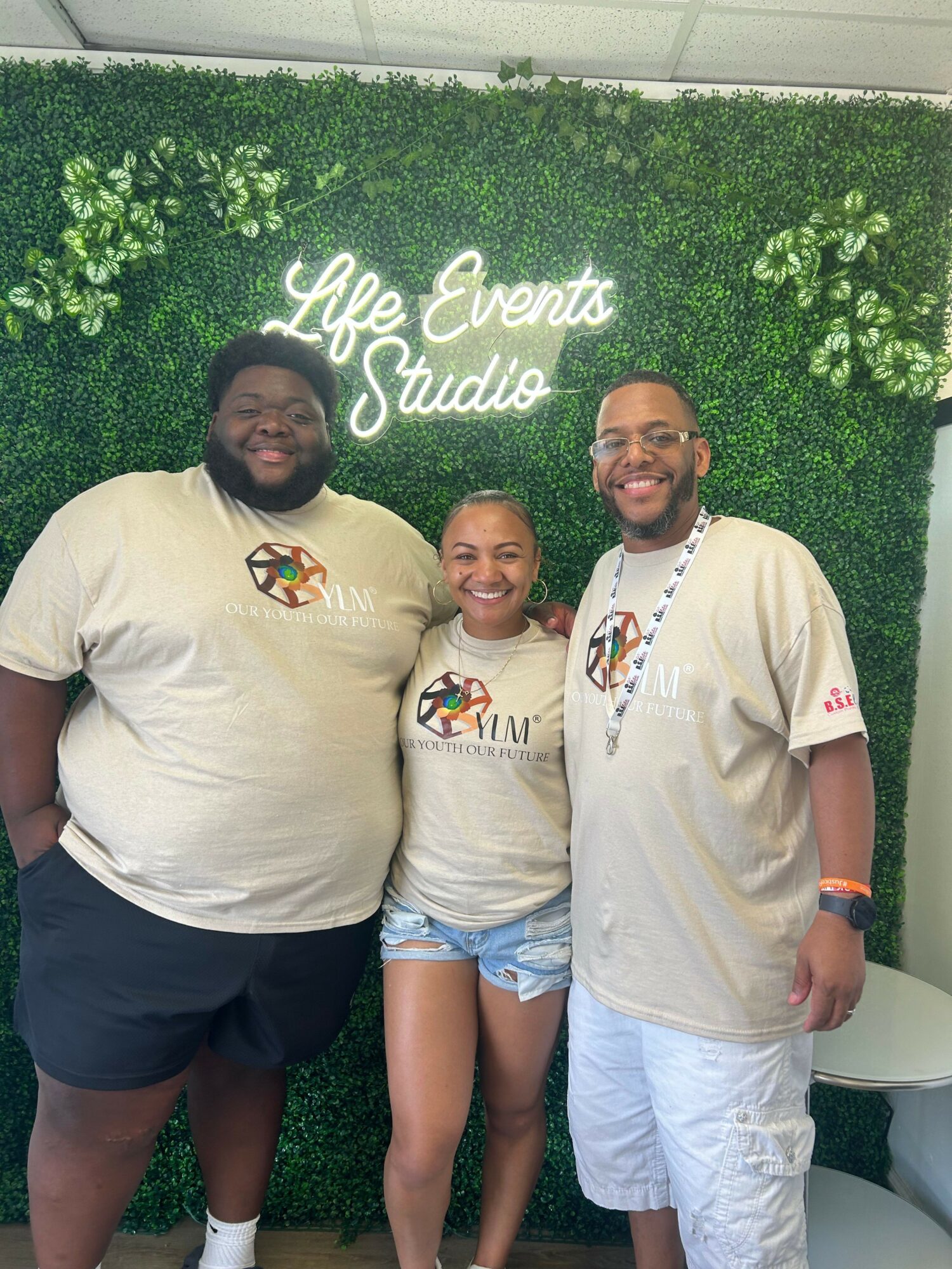 Three people standing in front of a green wall with white text, smiling, wearing matching beige shirts with a logo, and casual shorts.