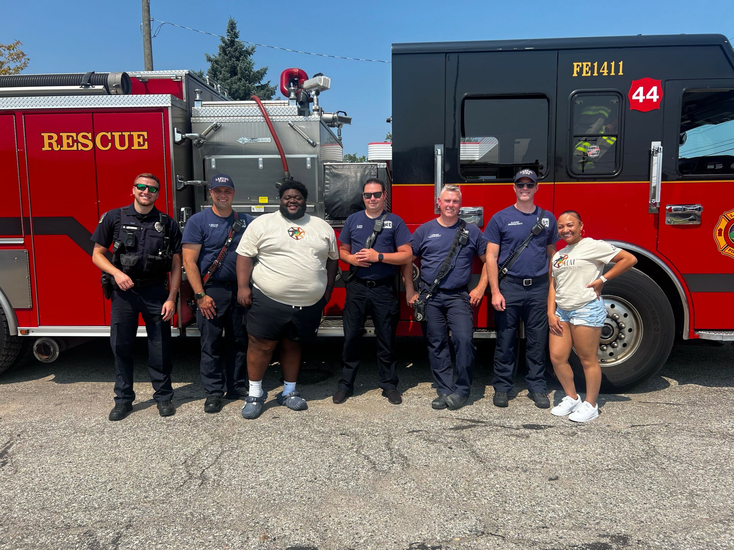 Group of people, including children and adults, standing on a fire truck, facing sideways, with a clear sky in the background.