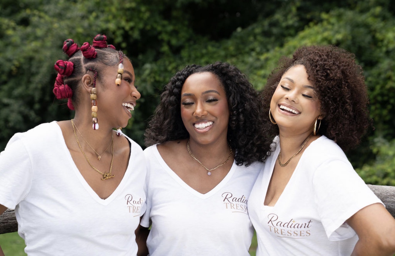 Three women smiling and laughing outdoors with green trees in the background, wearing white shirts with a logo.