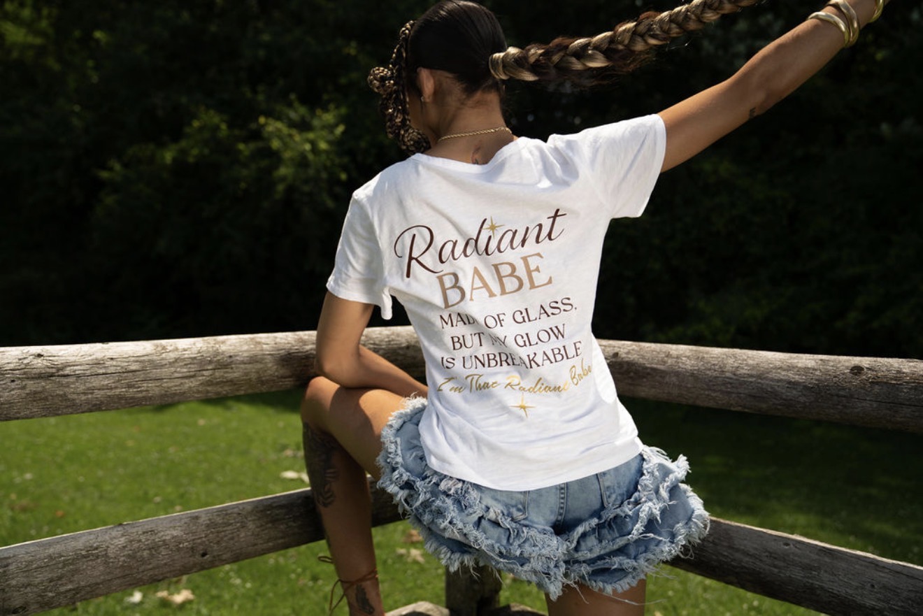 Person sitting on a wooden fence outdoors, wearing a white T-shirt and denim shorts, with trees in the background.