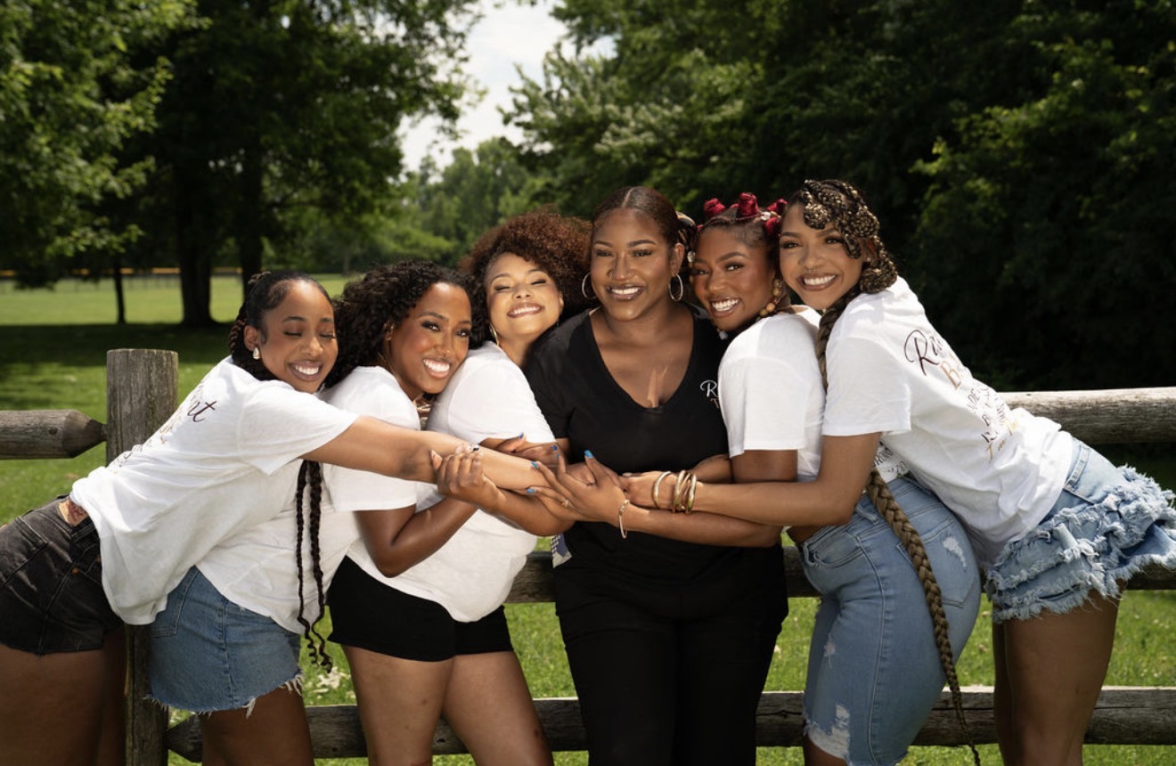 Group of six smiling women hugging outdoors with trees and a fence in background.