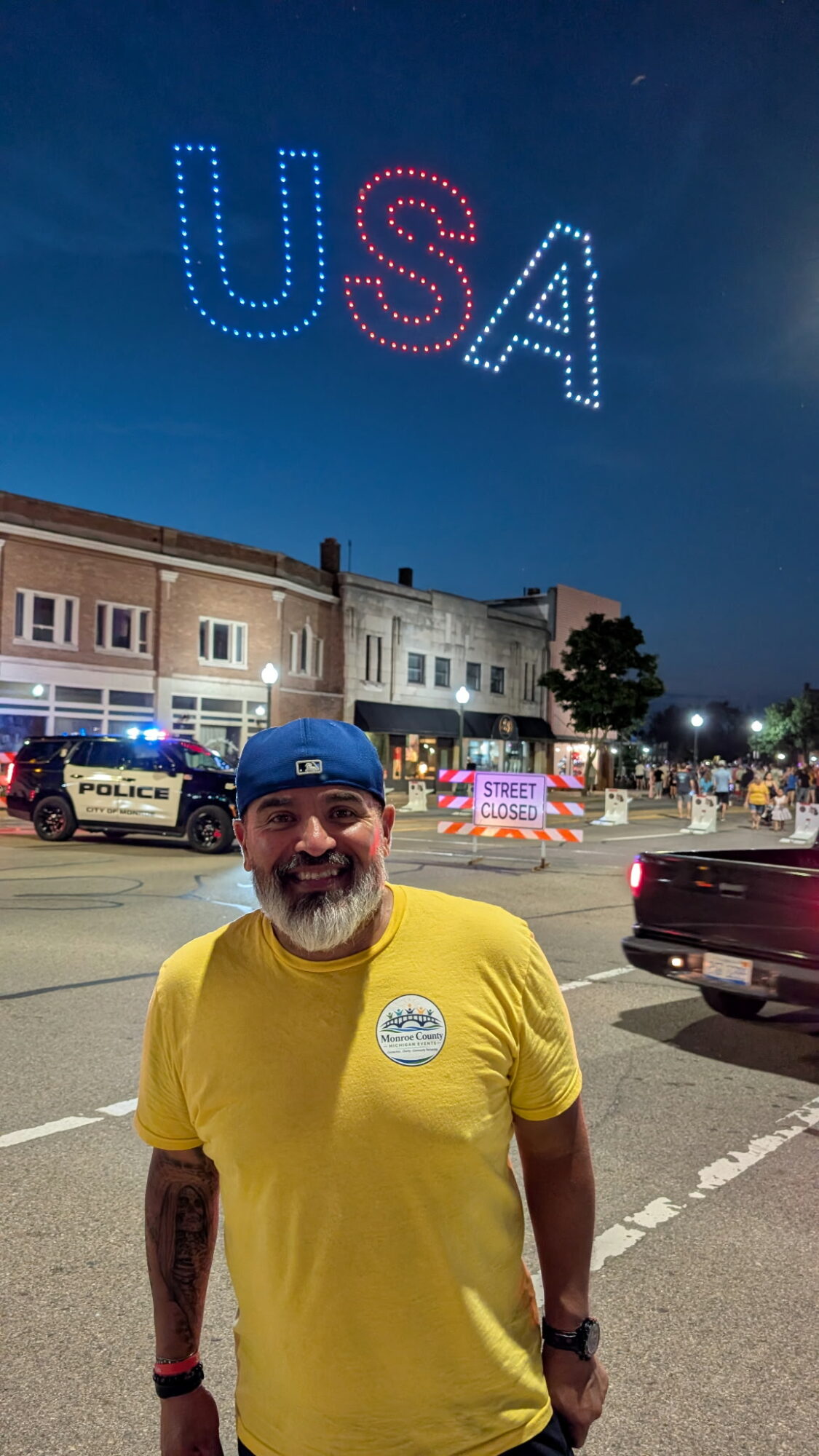 Man with a beard and blue cap smiling in a yellow shirt, standing on a city street at night with police cars and a large illuminated 'USA' sign in the sky.