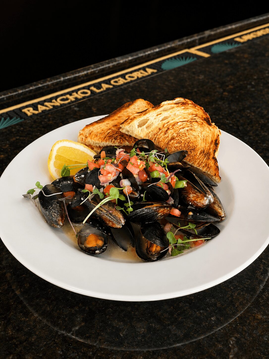 Mussels in broth with chopped herbs and diced tomatoes, served with toasted bread on a white plate.