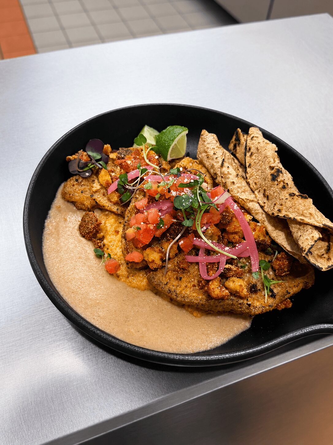 Black bowl with fried fish topped with salsa, lime, and pickled onions, served with tortilla chips and sauce, on a white surface.