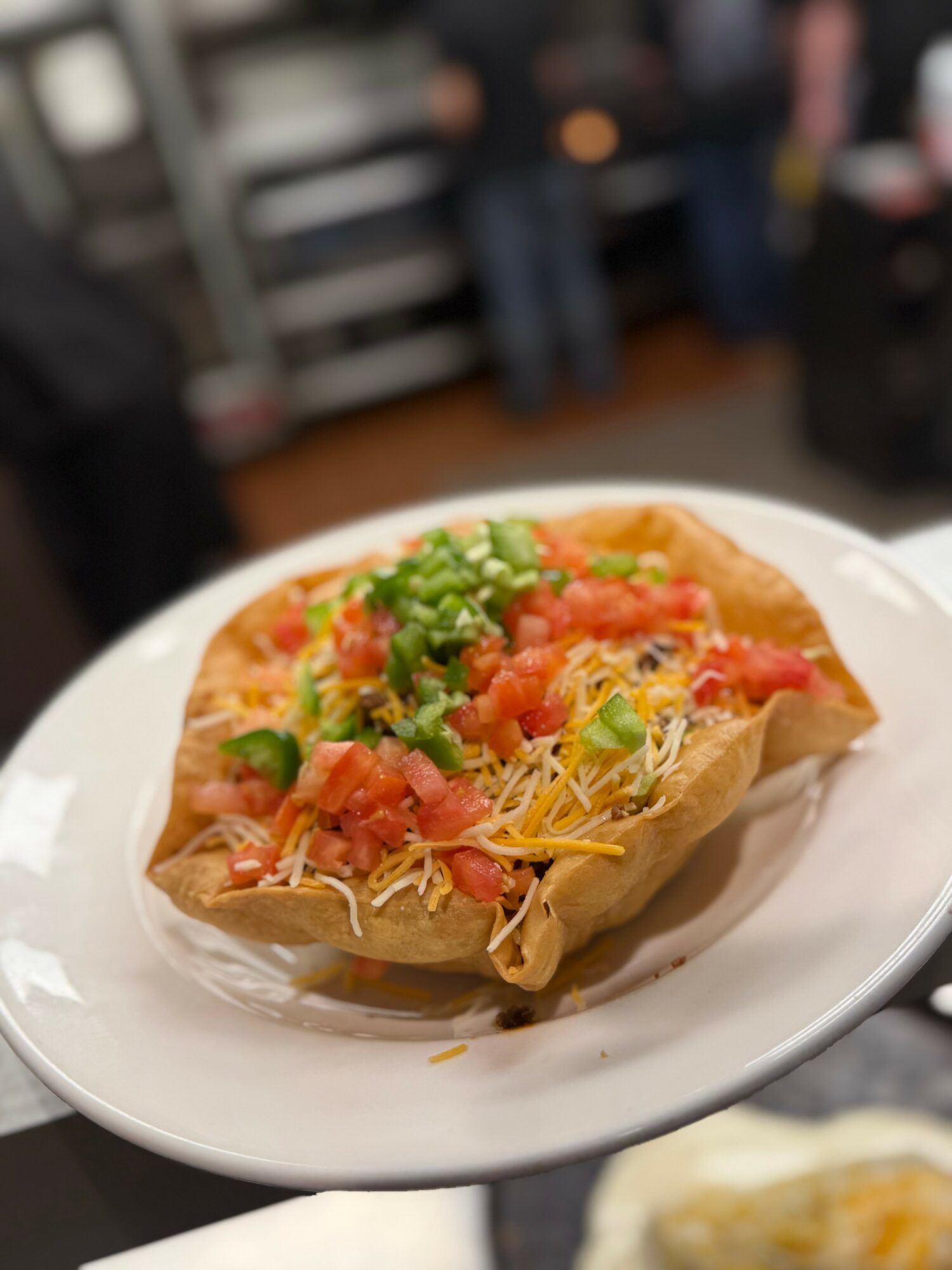 Taco with shredded cheese, chopped tomatoes, and green onions on a white plate, blurred background of a restaurant interior.