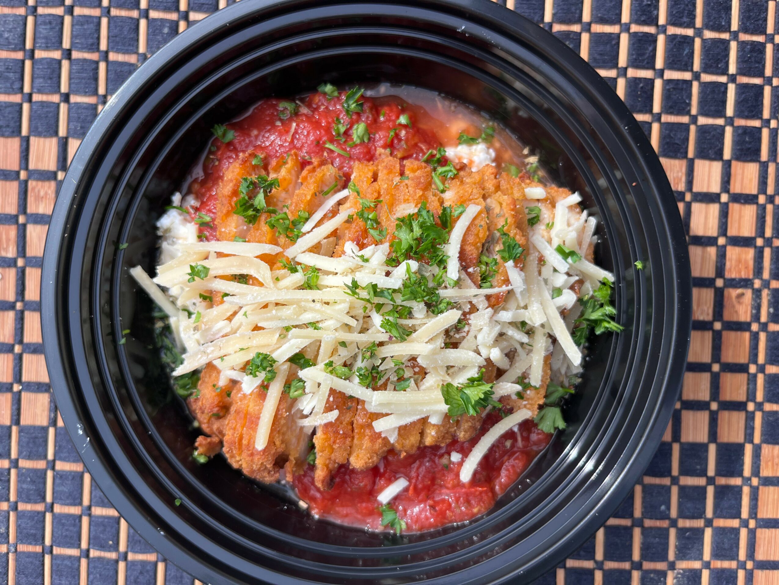Bowl of breaded fried fish topped with shredded cheese and chopped herbs, with tomato sauce underneath, on a woven mat