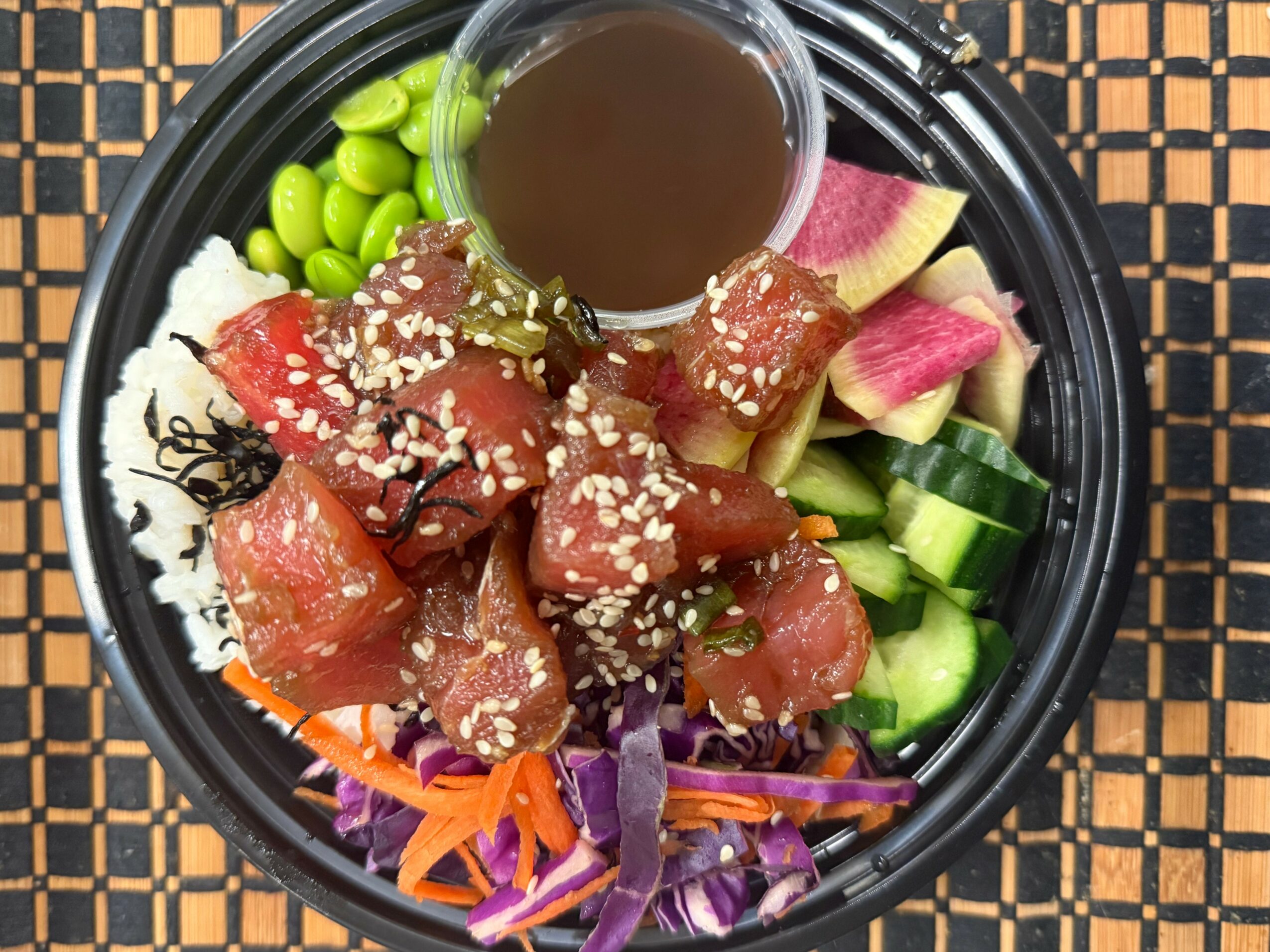 Bowl of raw fish, vegetables, and sauce with sesame seeds on a black plate, on a woven mat.