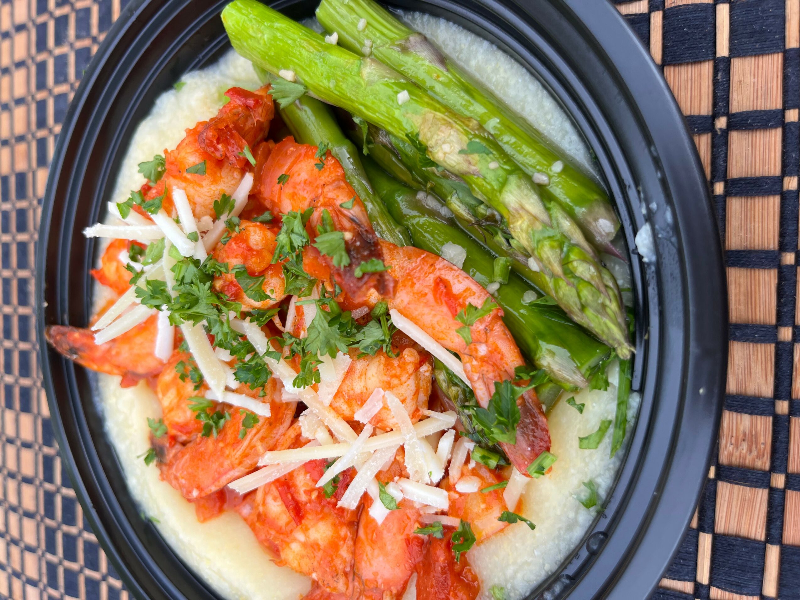 Shrimp with green asparagus on a bed of creamy sauce, garnished with shredded cheese and herbs, in a black bowl.