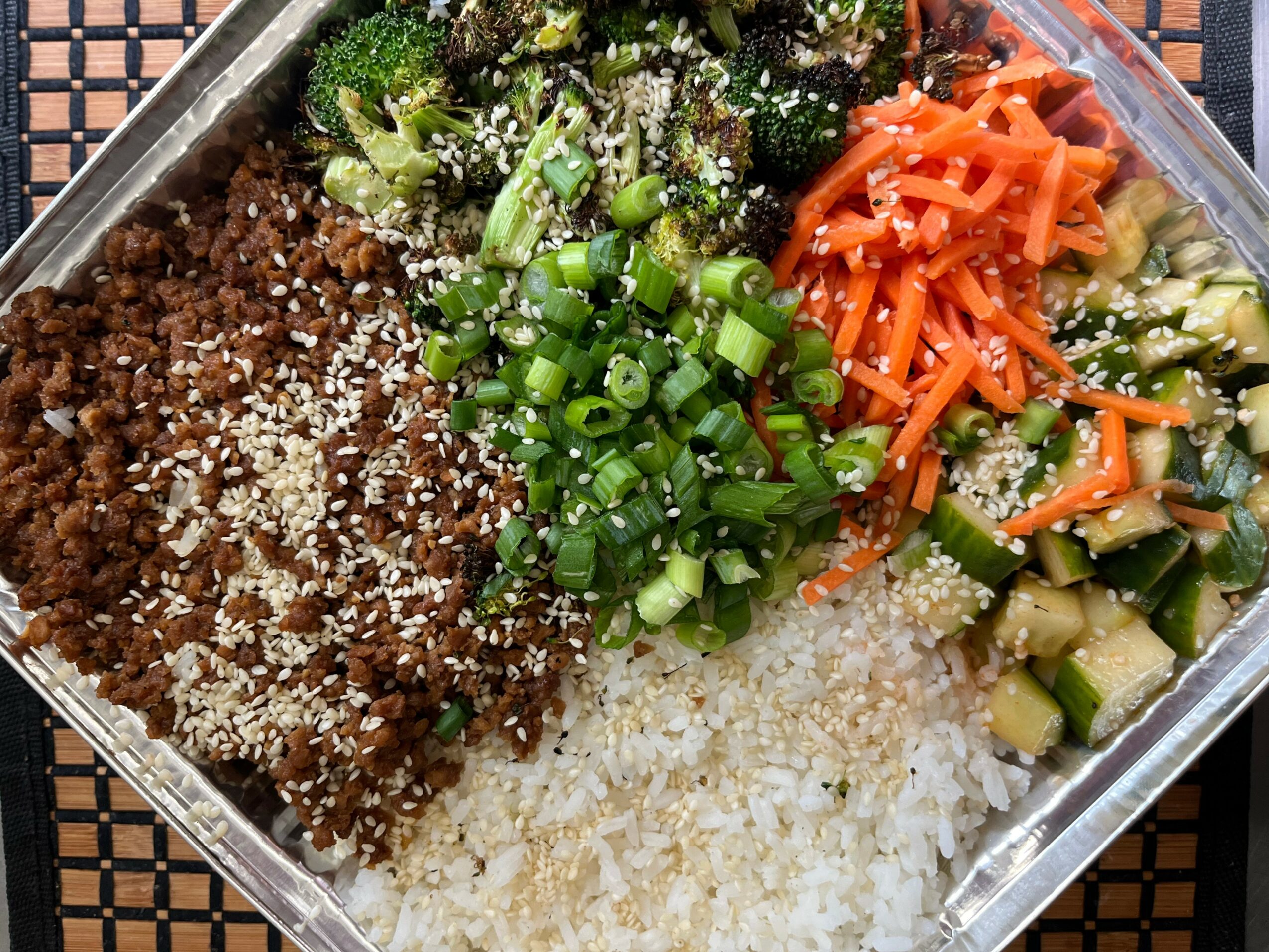 Assorted vegetables and grains in a container, including chopped green onions, shredded carrots, cucumber, and cooked grains.
