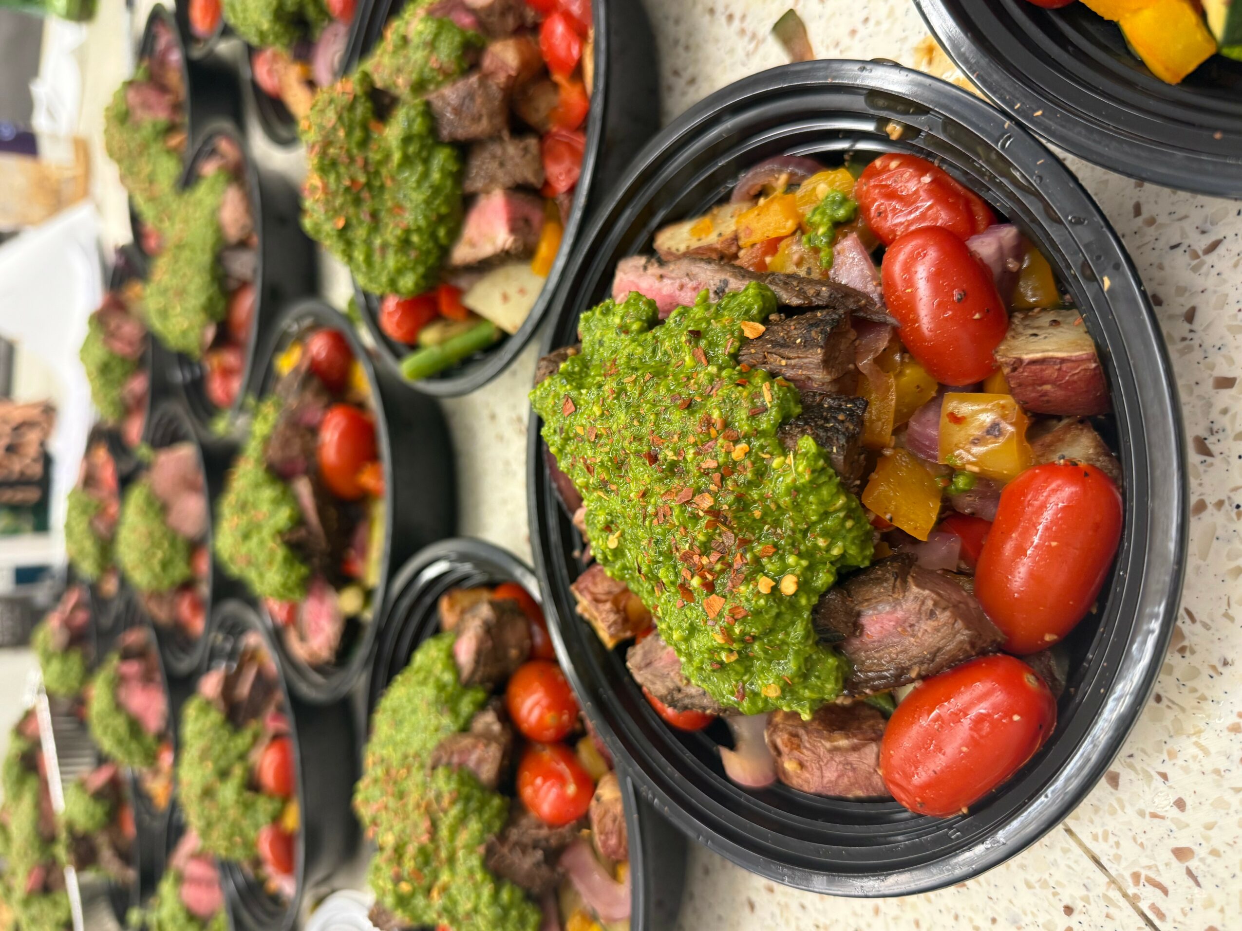 Black plastic container with cooked vegetables including cherry tomatoes, broccoli, and yellow bell peppers, on a gray surface.