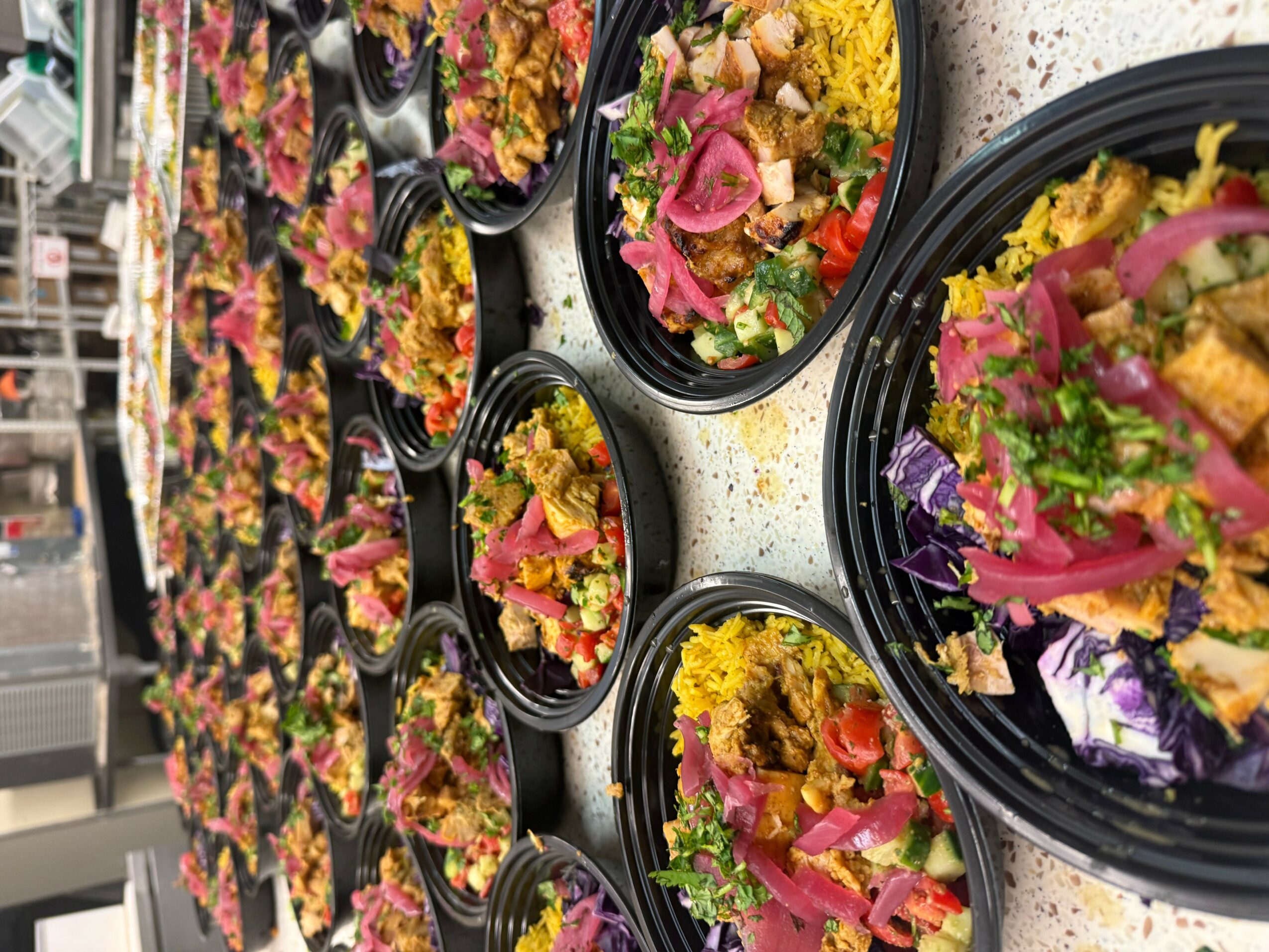 Multiple black bowls filled with colorful salads on a table, with a background of more salads and utensils.