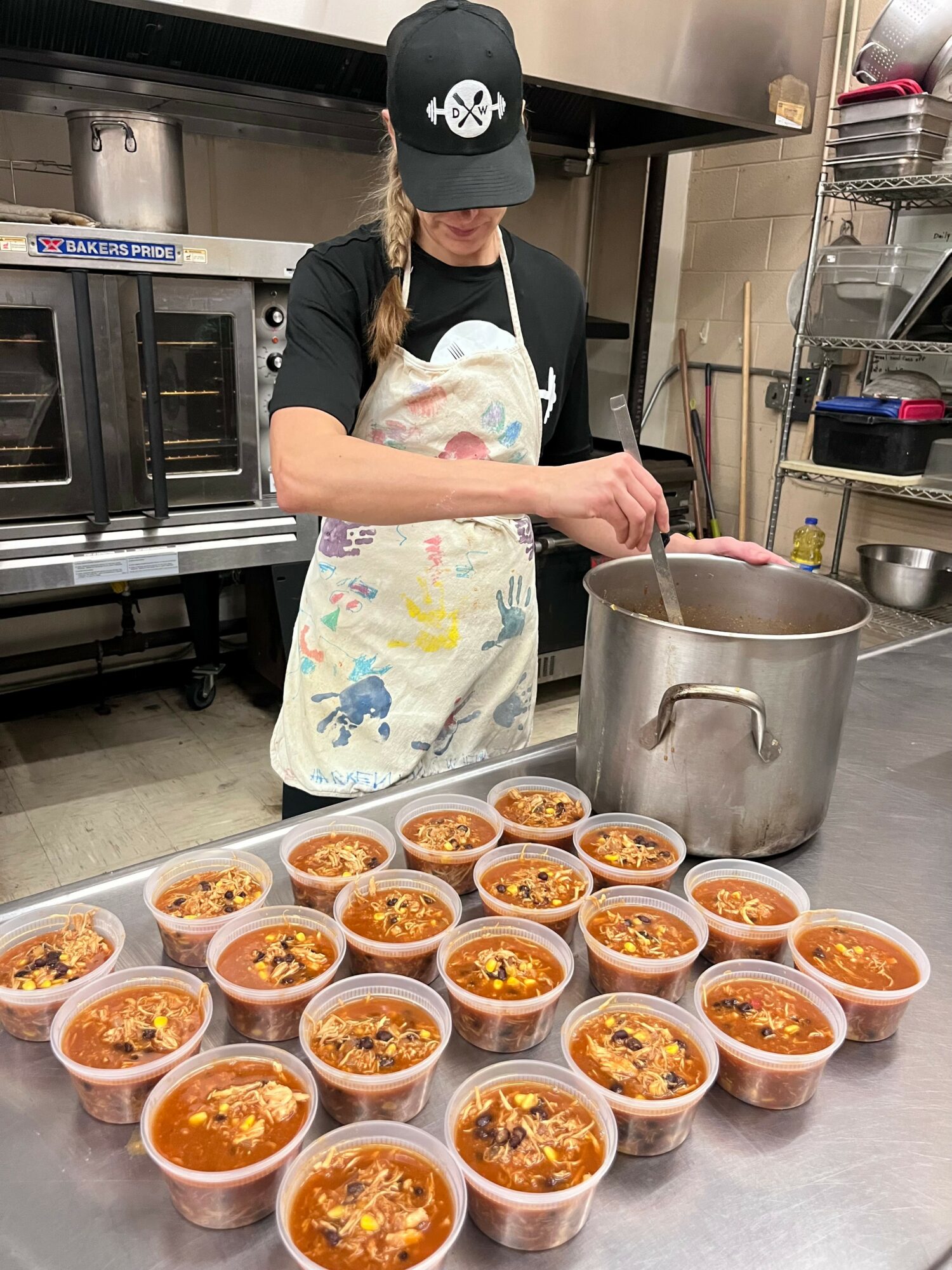 Person wearing a black cap and apron preparing food in a kitchen with bowls of soup or stew on the counter.