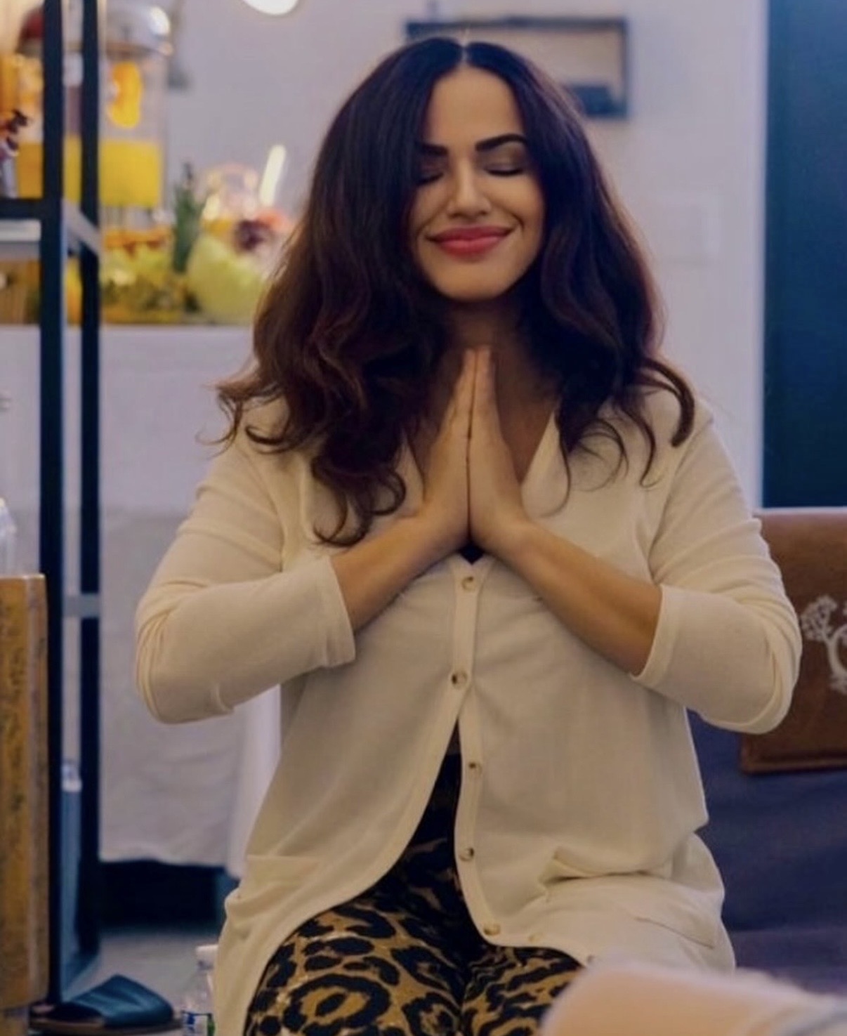 Woman with long dark hair and closed eyes, smiling with hands pressed together in a prayer gesture, sitting indoors.
