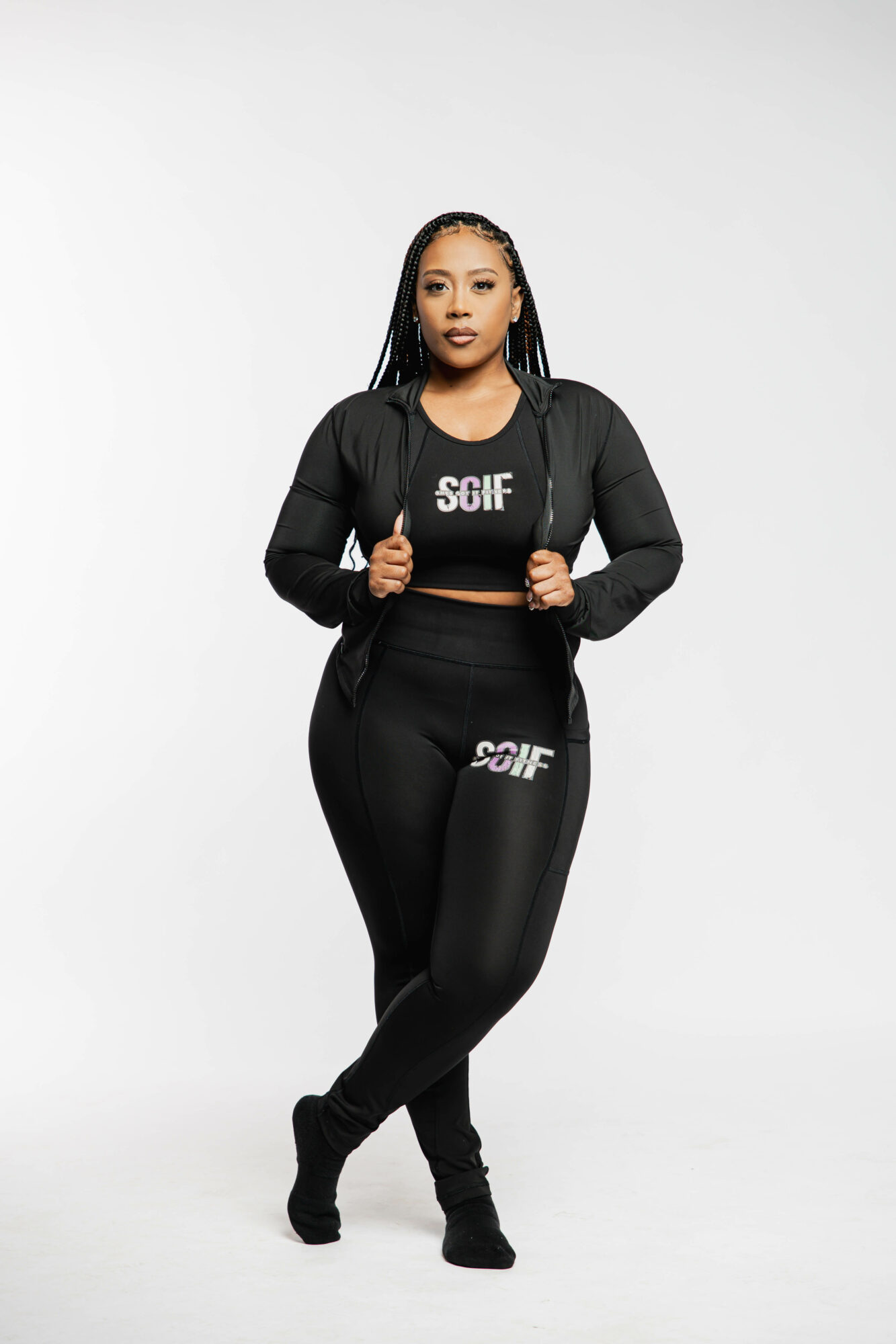 Woman in black athletic outfit standing with hands on jacket, plain white background, confident expression.