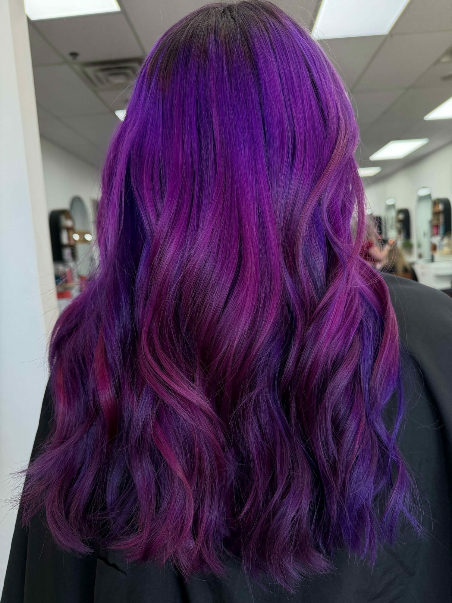 Person with long, wavy hair dyed in shades of purple and pink, viewed from the back in an indoor setting.