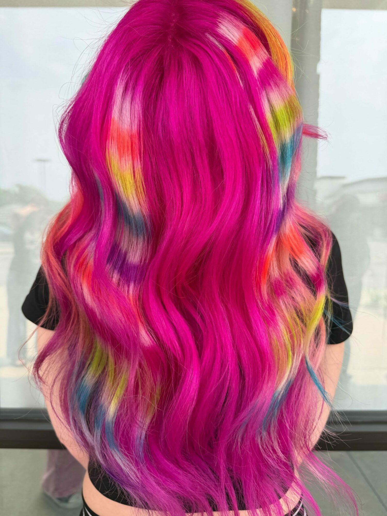Person with long, wavy hair dyed bright pink with streaks of yellow and orange, viewed from behind.