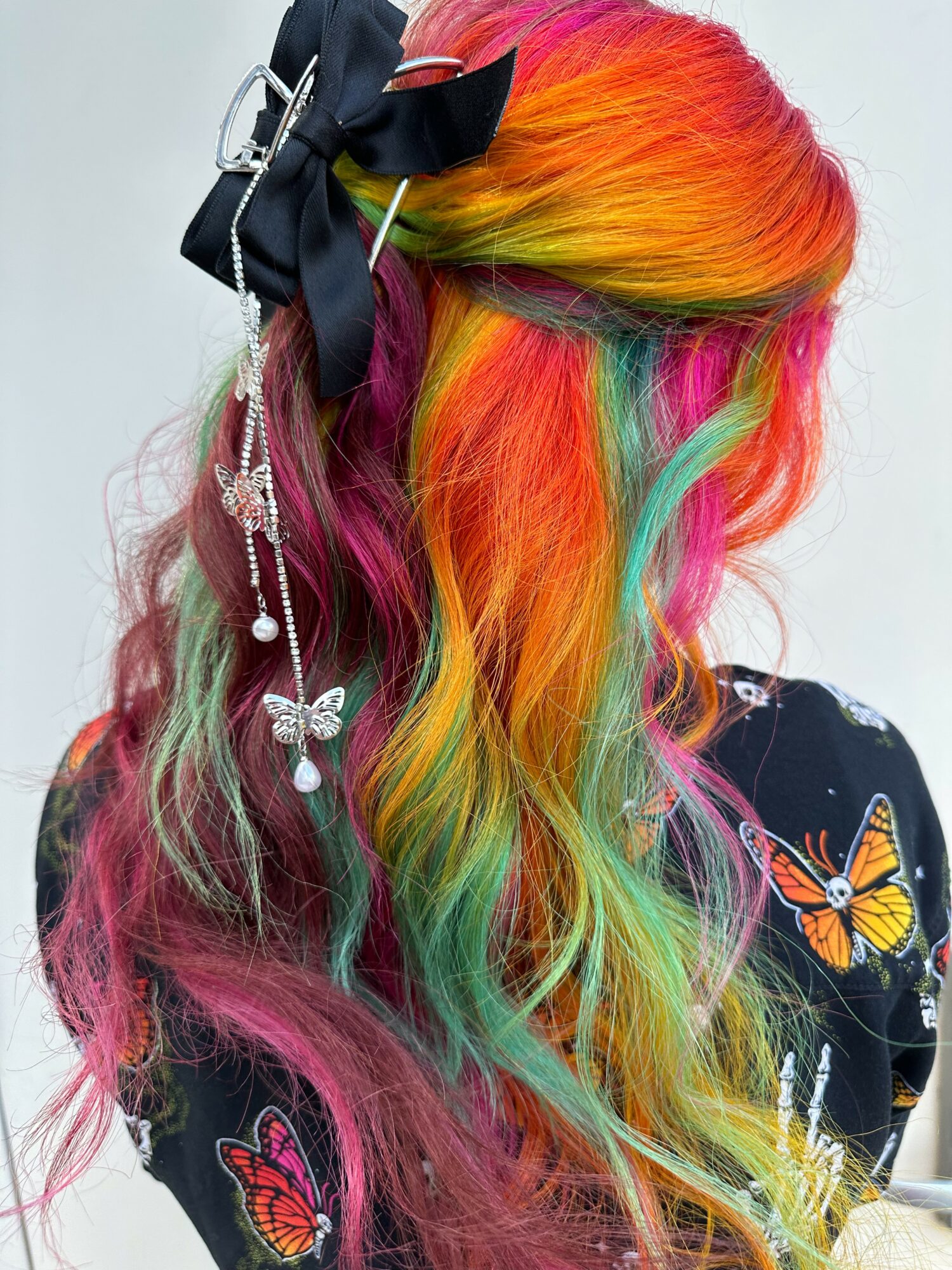 Person with long, rainbow-colored hair styled with a black bow and decorative accessories, wearing a butterfly-patterned top.