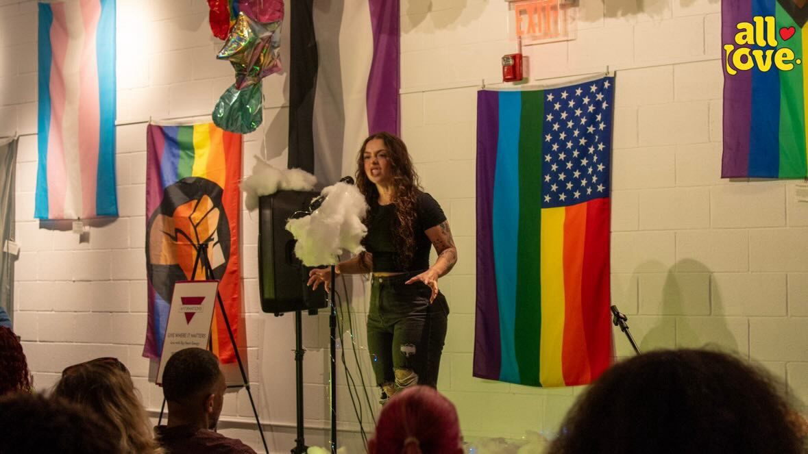 Person standing on stage with rainbow flags and a cloud of smoke, audience watching, colorful decorations, pride event.
