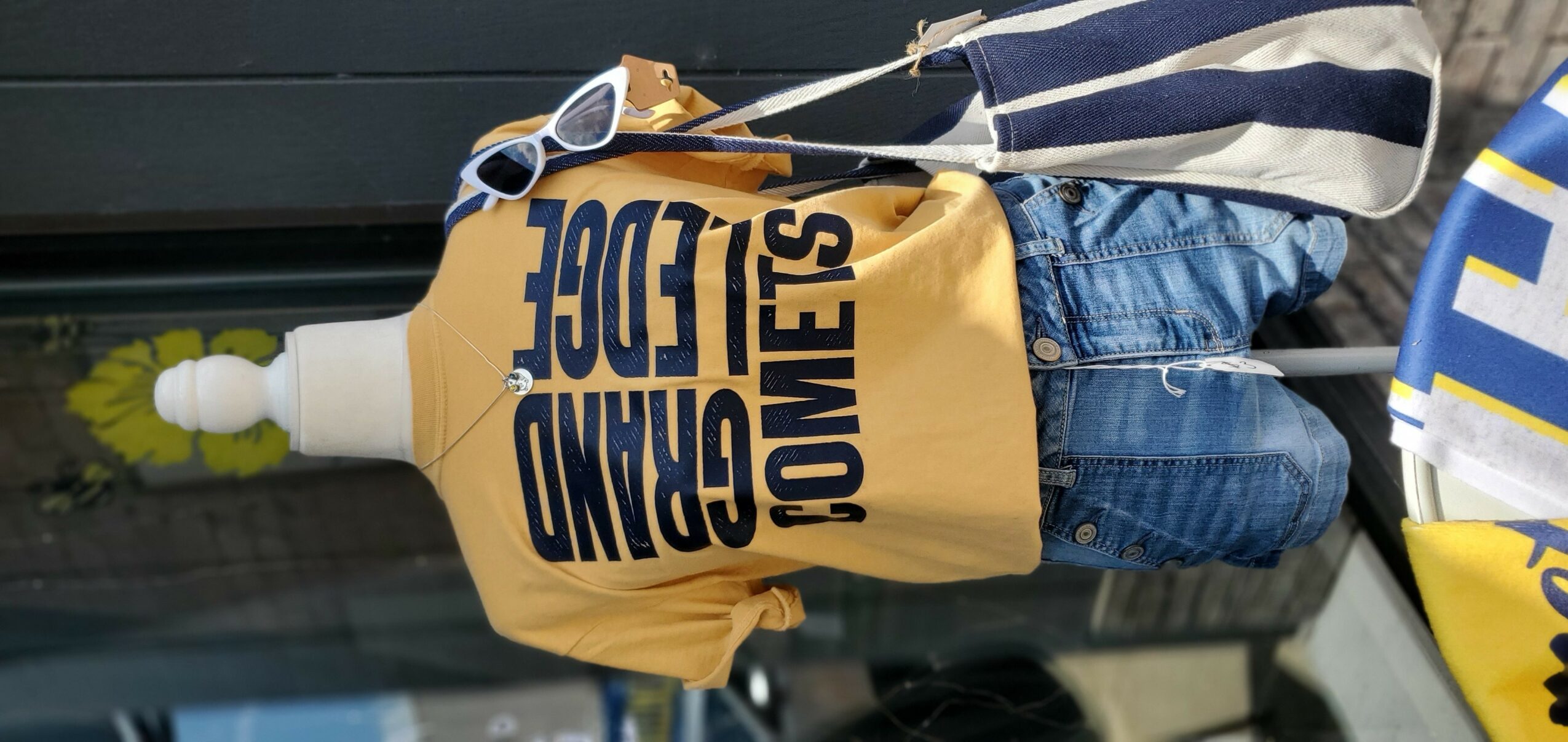 Mannequin wearing a yellow T-shirt with black text, blue jeans, and holding a striped bag, standing outdoors.