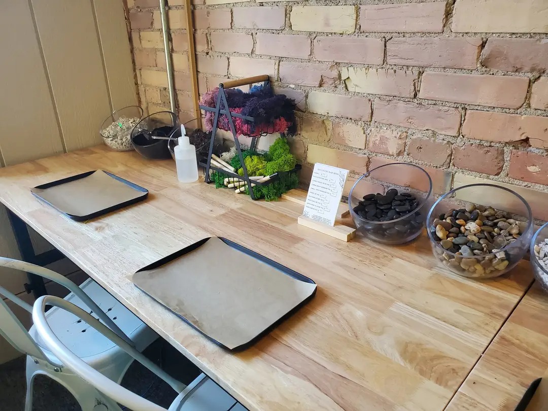 Wooden table with trays, containers of stones, a plant, and a small sign, against a brick wall background.