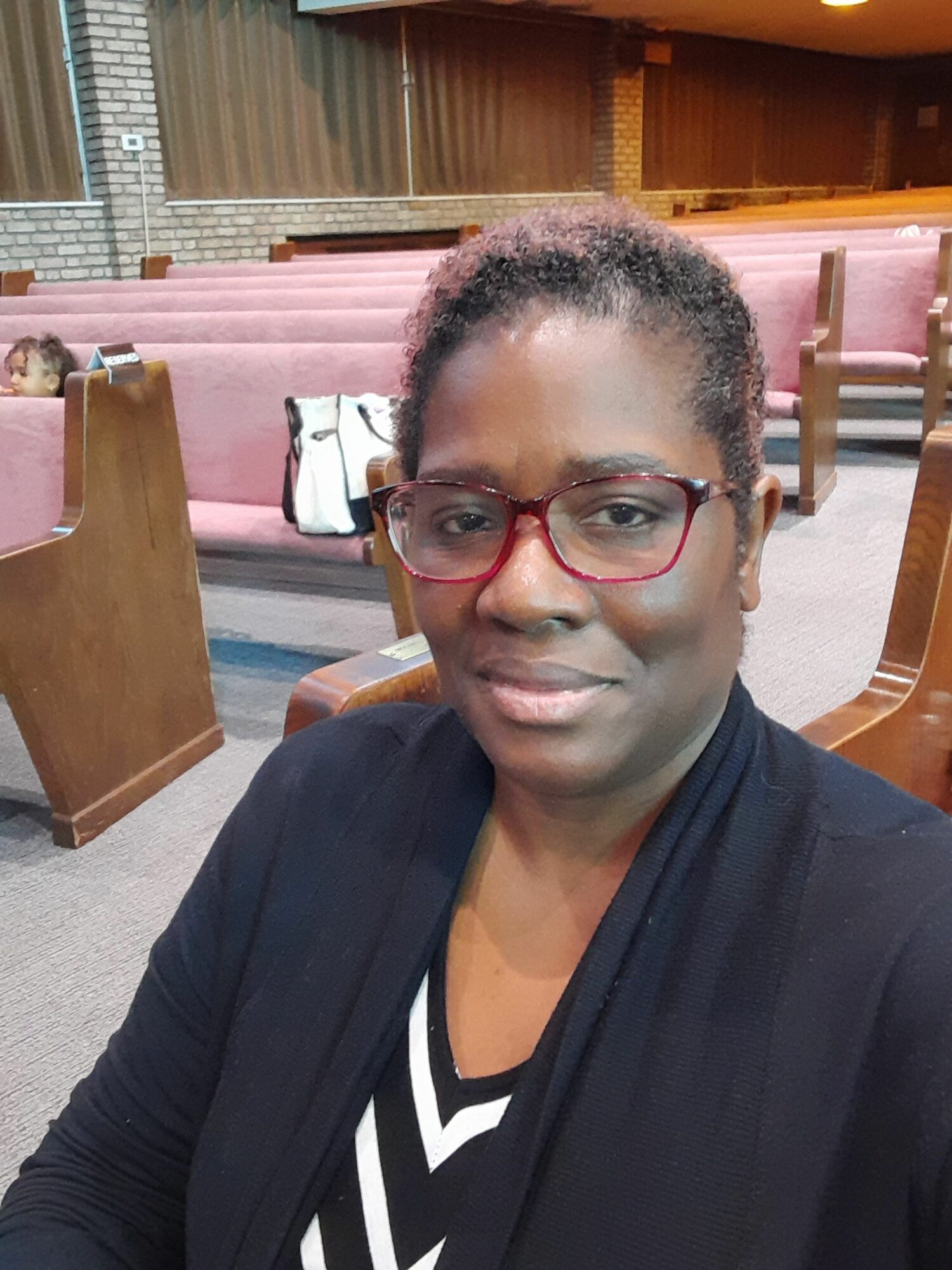 Woman with glasses and curly hair sitting indoors, background with pink seating and wooden furniture.