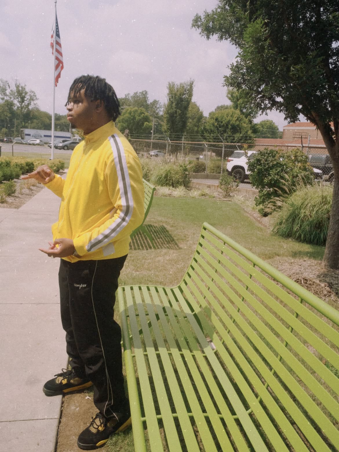Young man in yellow jacket standing next to a green bench outdoors, with trees and a flag in the background.