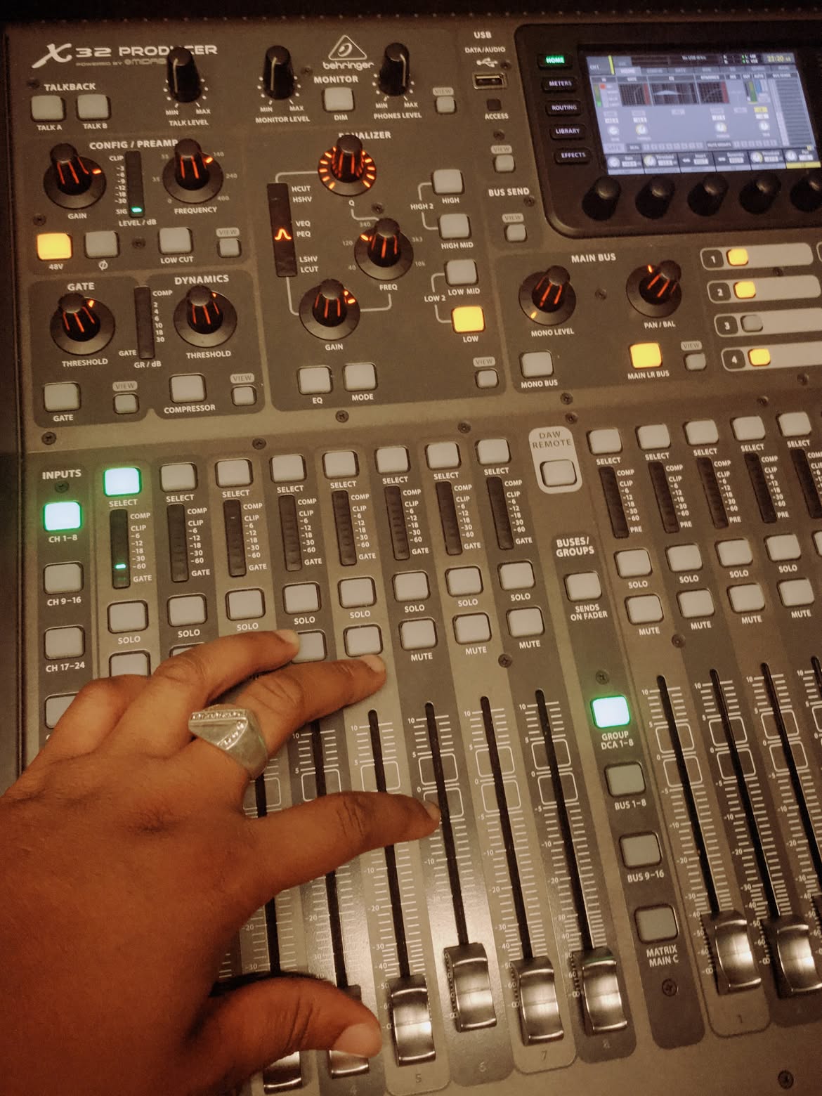 Person's hand adjusting sliders on a music mixing console with knobs and a screen, viewed from above.