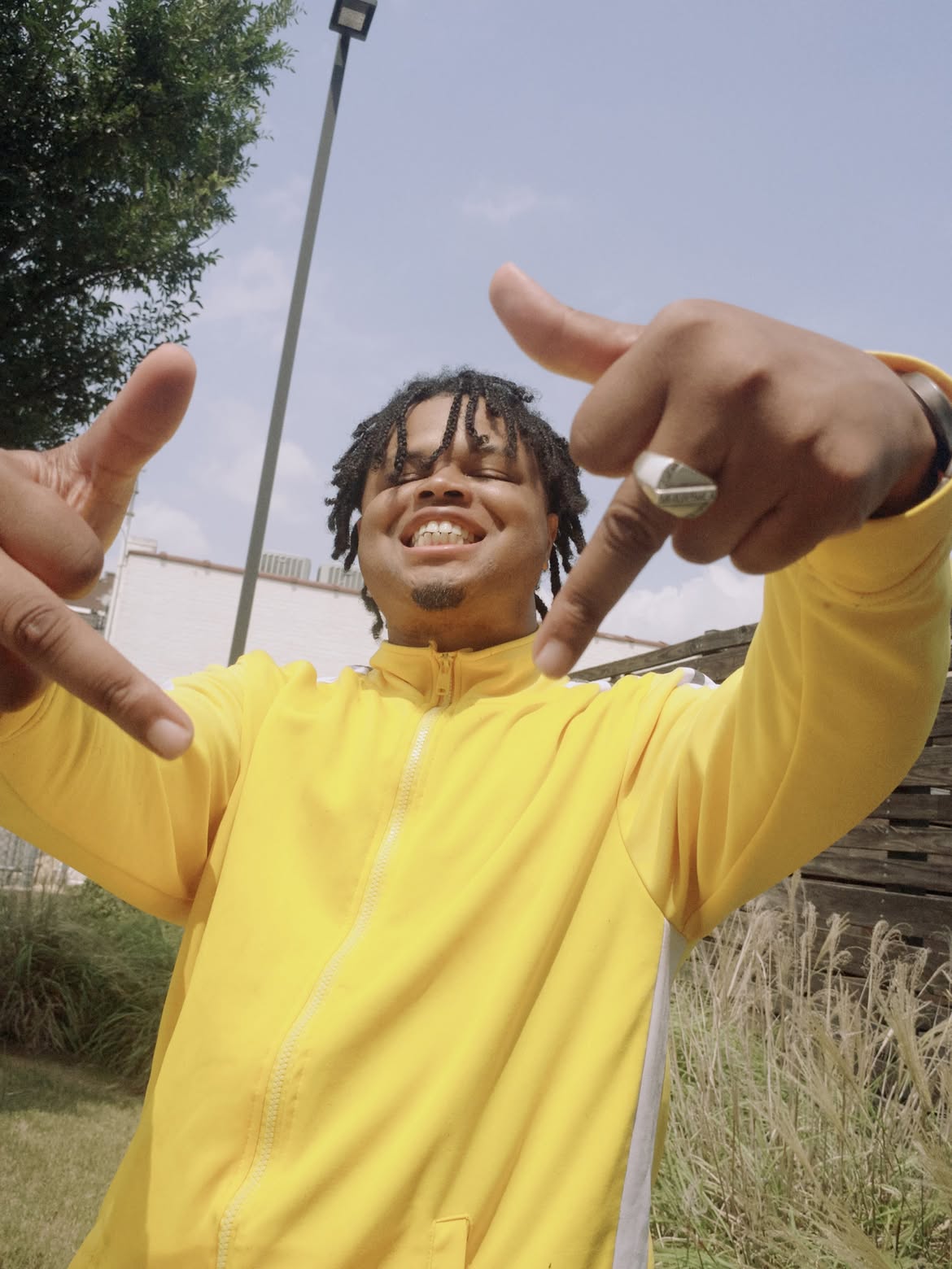 Young man with dreadlocks smiling and making hand gestures outdoors, wearing a yellow jacket.
