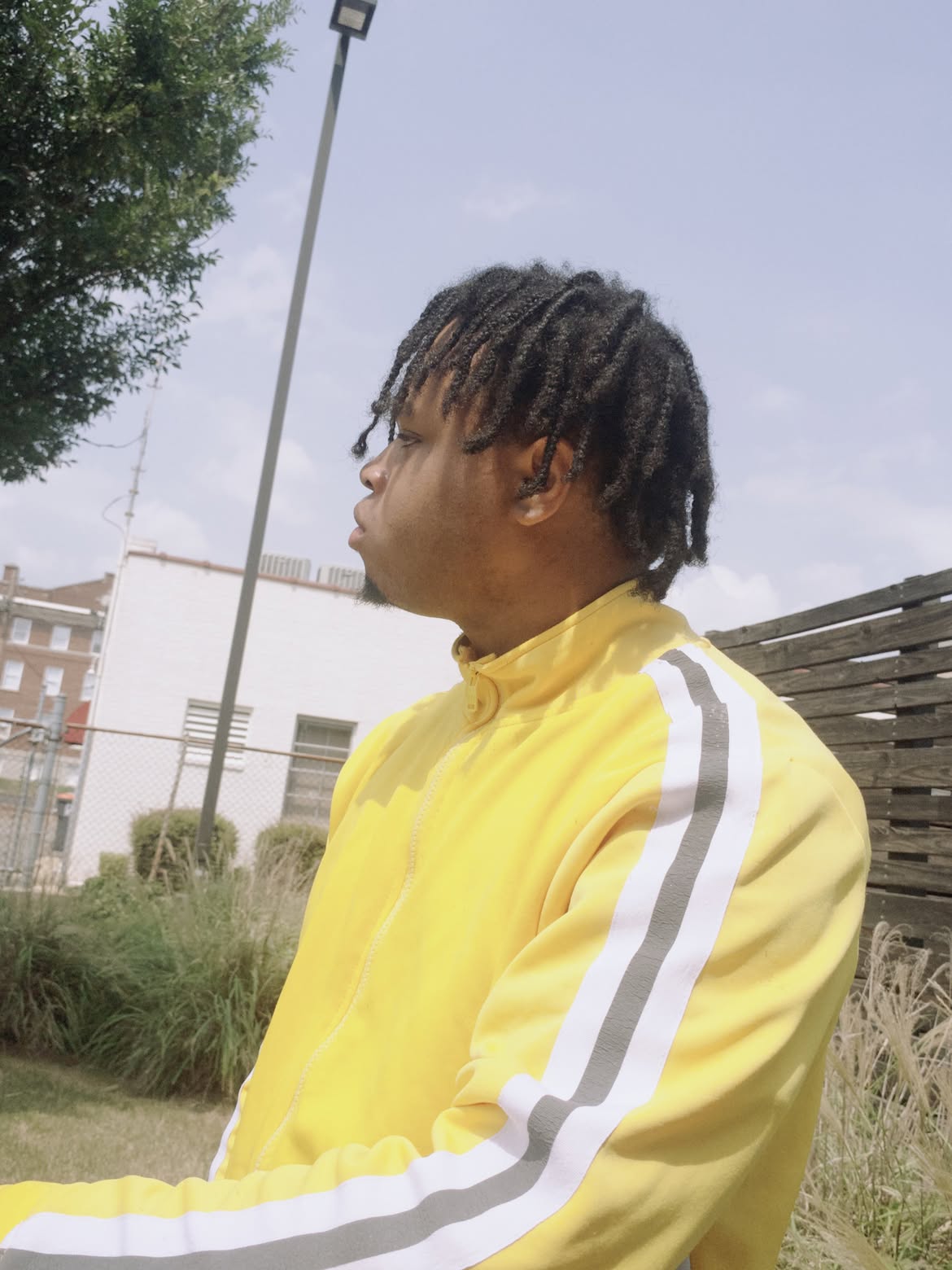 Person with dreadlocks in a yellow jacket outdoors, facing left, with a fence and buildings in the background.