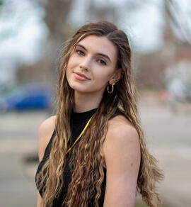 Young woman with long wavy hair, wearing a black sleeveless top and hoop earrings, outdoors with blurred background.