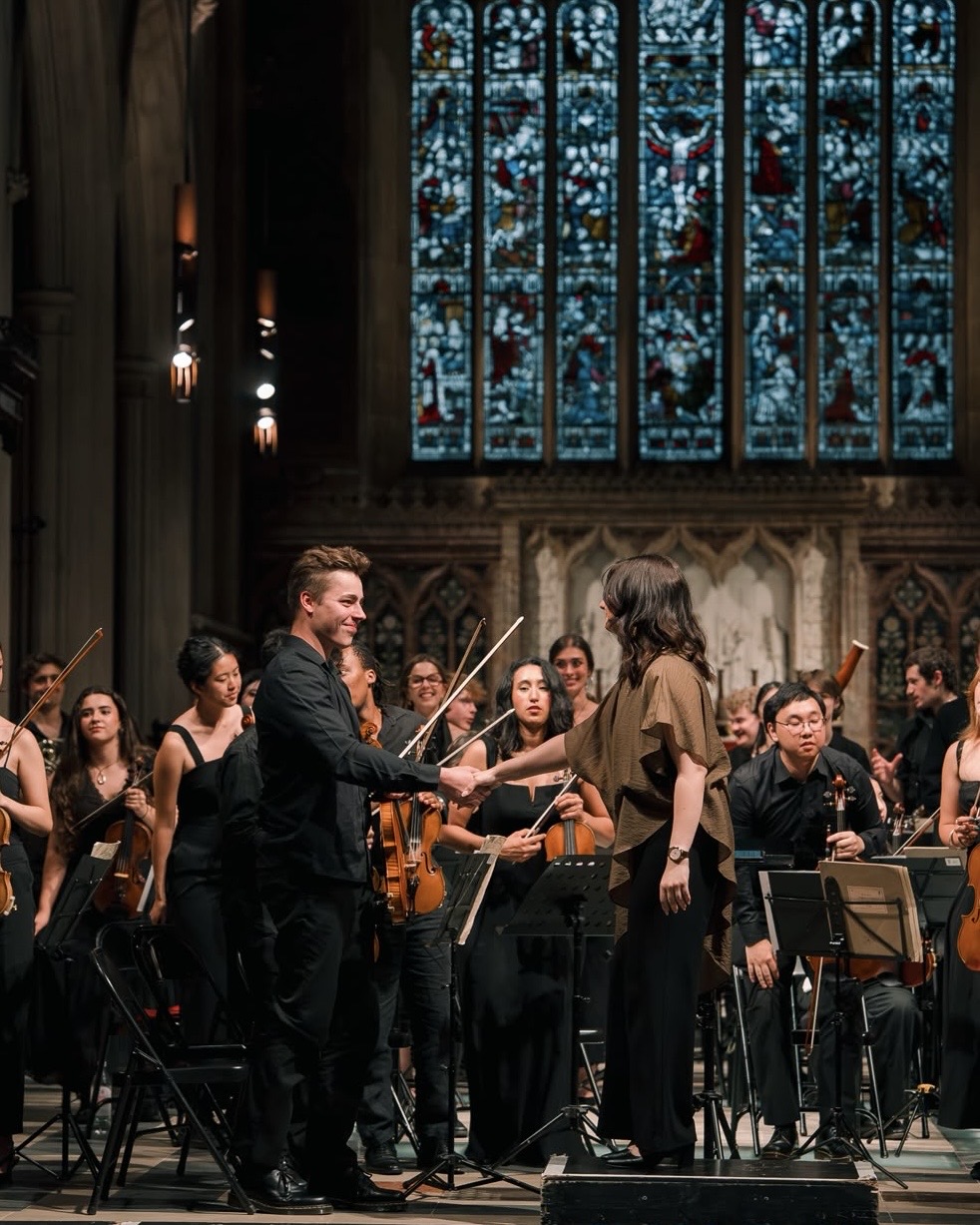 Orchestra performing in a church with stained glass windows, conductor and musicians playing instruments, audience in background.