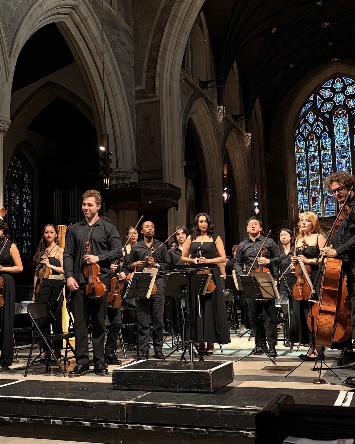 Orchestra standing on stage in a cathedral with stained glass windows, holding instruments, after performance or rehearsal.