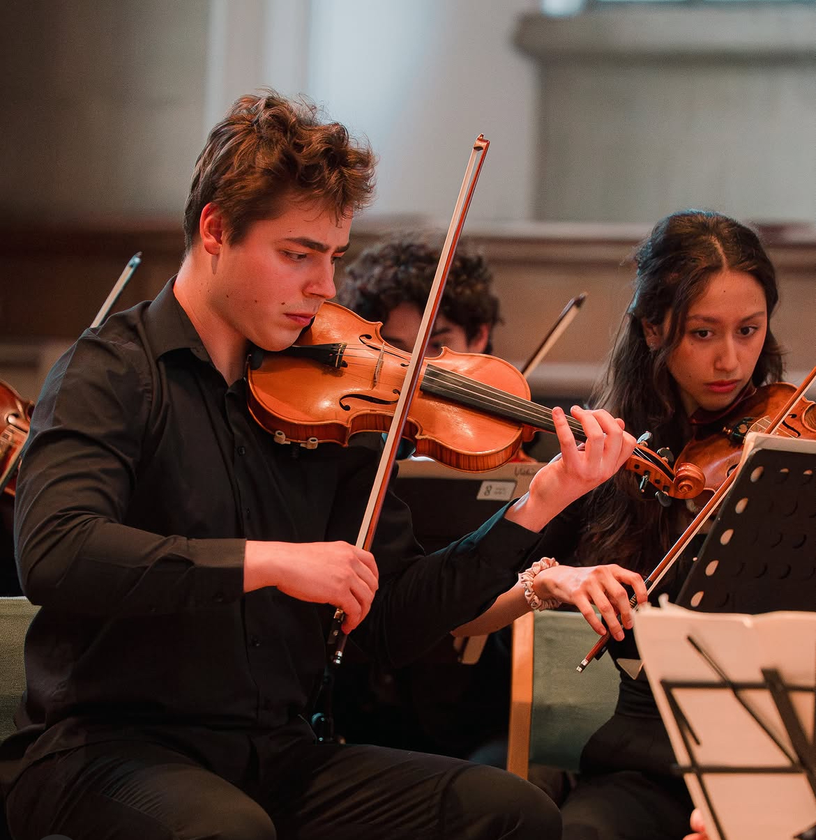 Young man playing violin in an orchestra, with other musicians visible in the background.