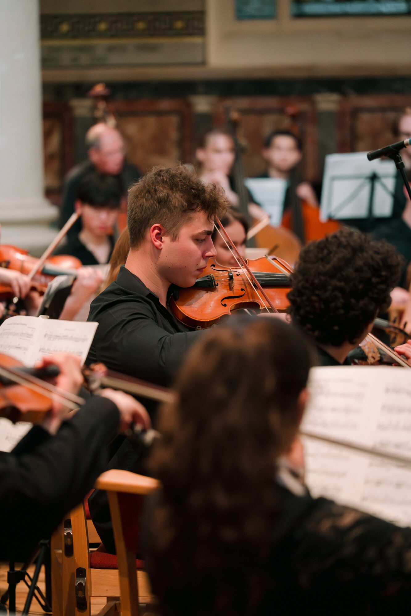 Young musician playing violin in an orchestra with other musicians and sheet music around him.