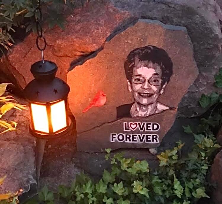 A painted portrait of a smiling person with glasses on a rock, surrounded by greenery and a lantern, with a 'Loved Forever' sign.