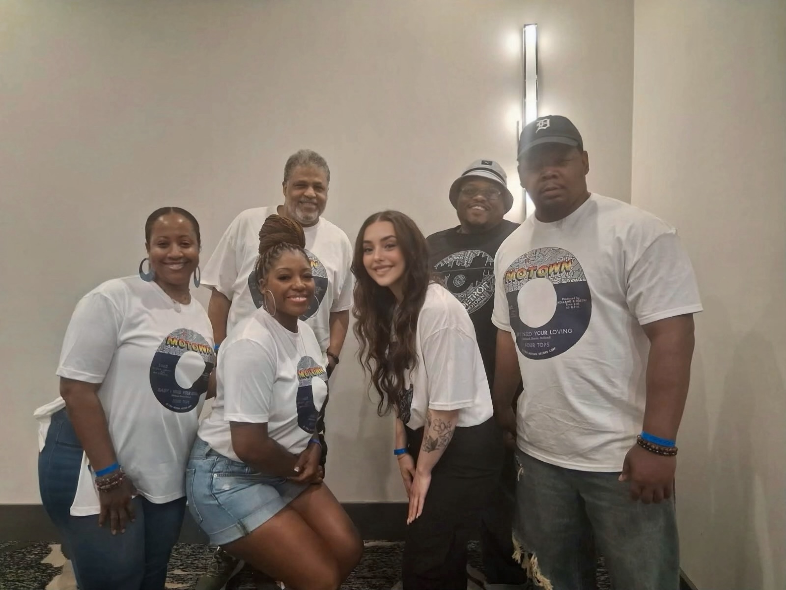 Group of six people standing indoors, smiling, wearing matching white T-shirts with a circular logo, posing for a photo.