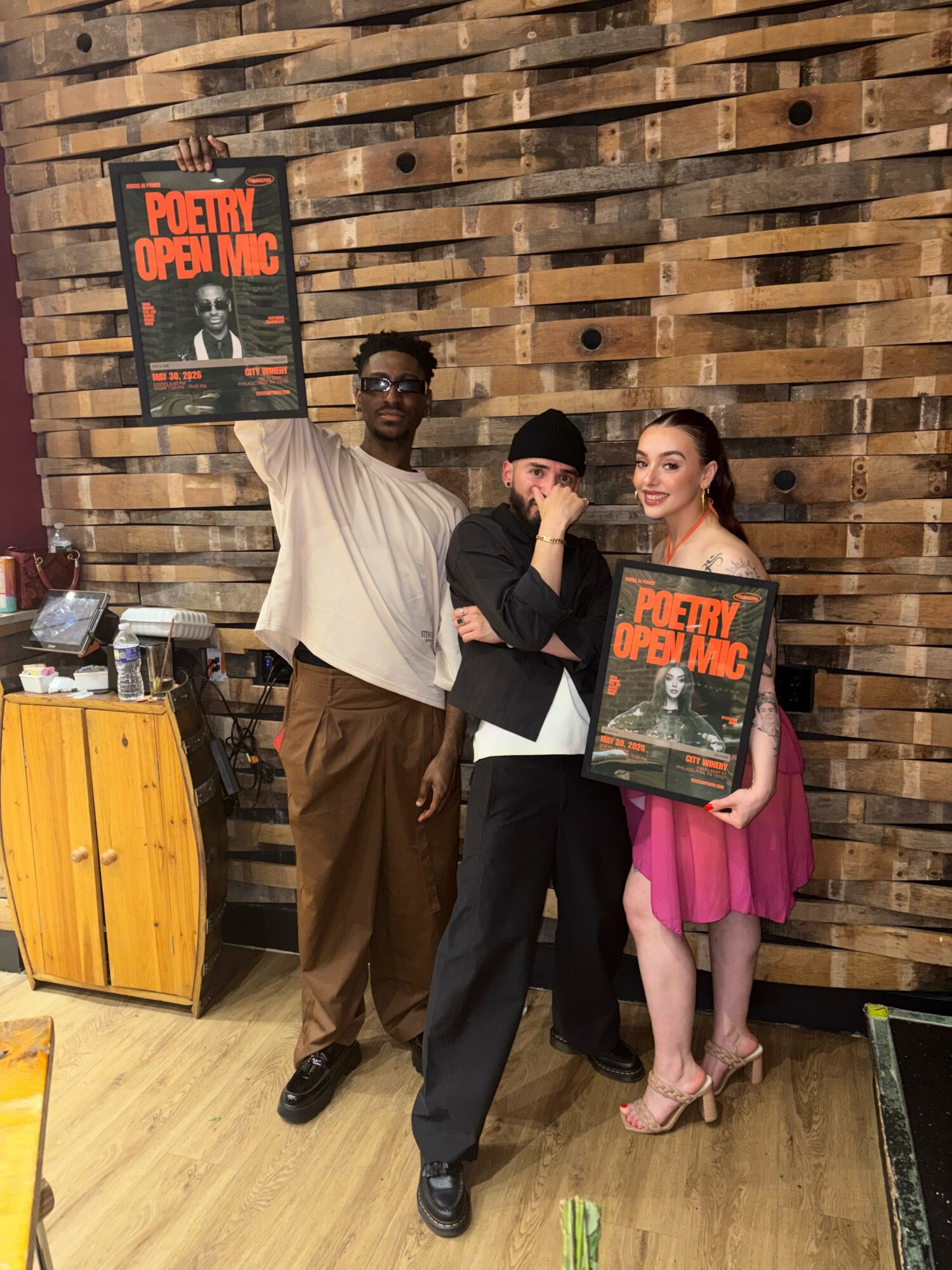 Three people standing indoors against a wooden wall, holding posters for poetry open mic event, with a yellow cabinet nearby.